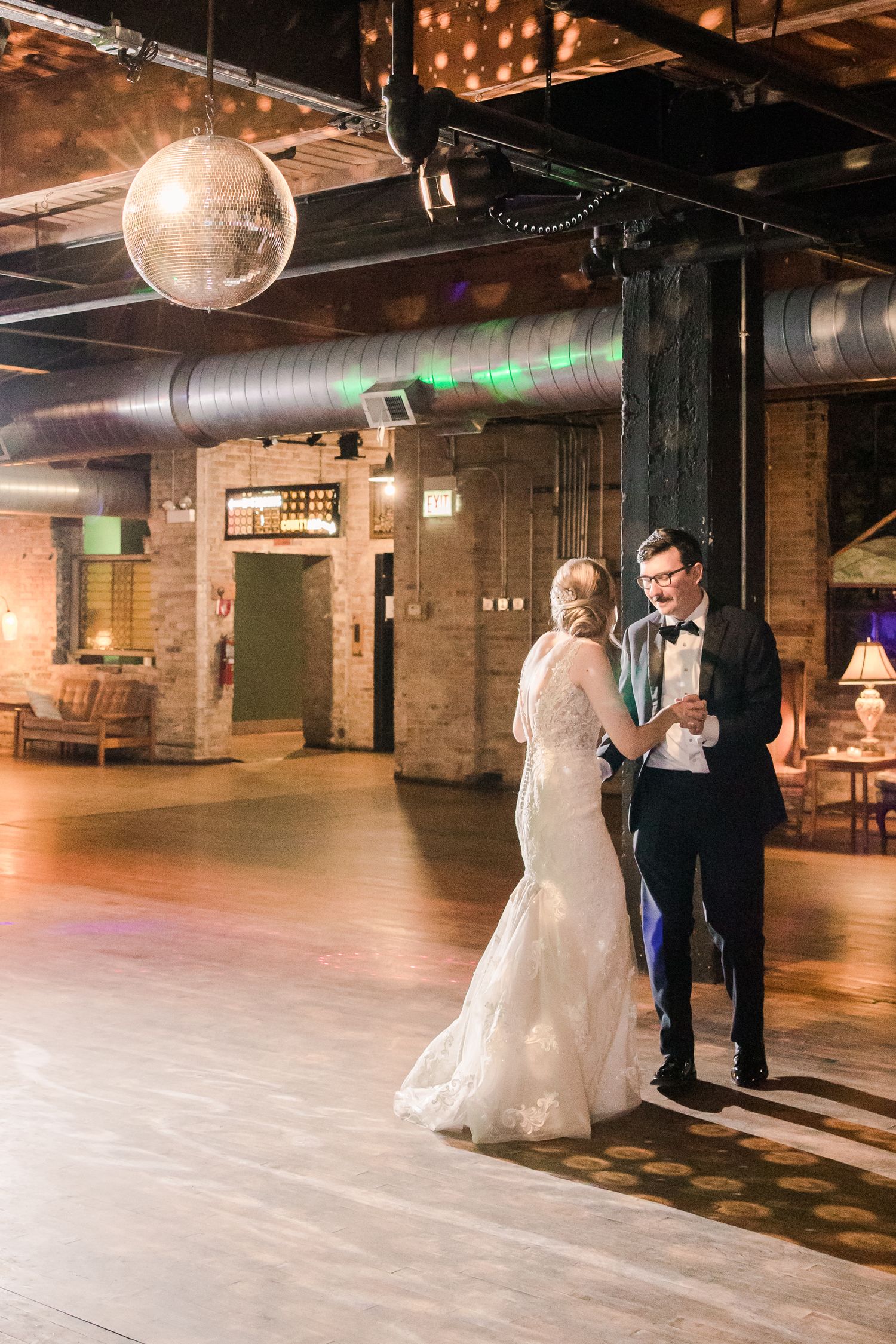 Couple sharing intimate moment under disco ball in industrial-style venue with exposed brick walls.