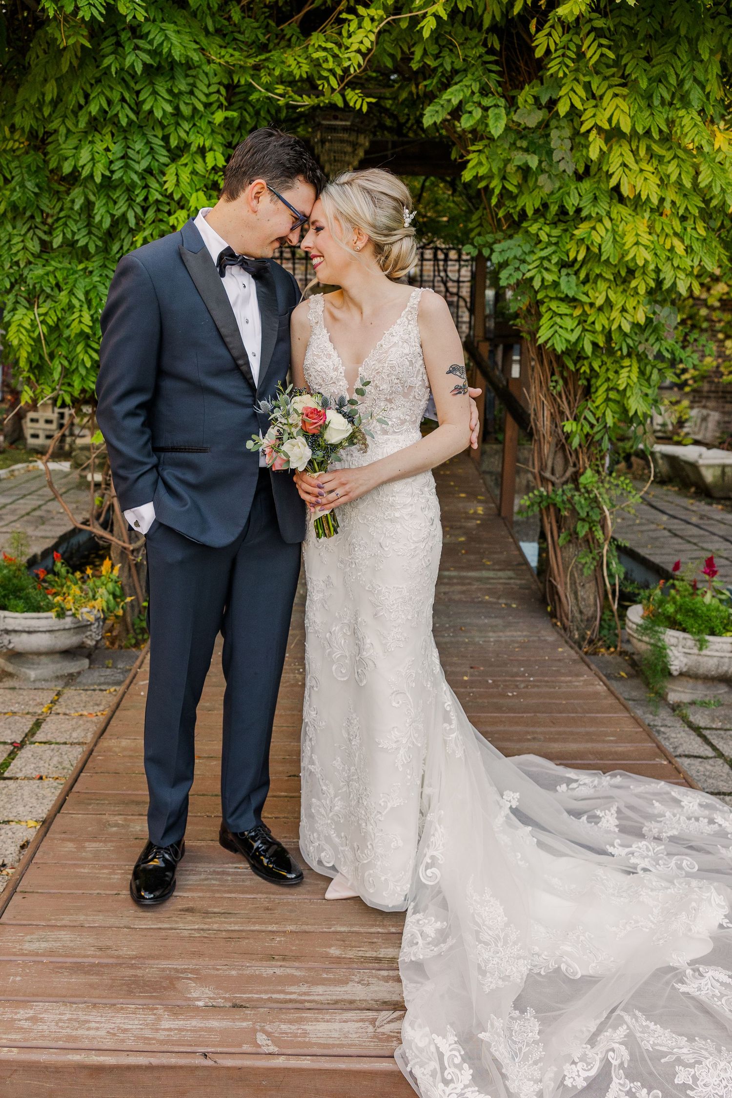 Romantic wedding portrait on wooden path with lush greenery backdrop and flowing bridal train.