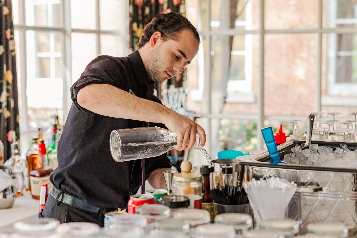 Bartender carefully pouring a cocktail at an elegant bar setup with various bottles and supplies in the background.