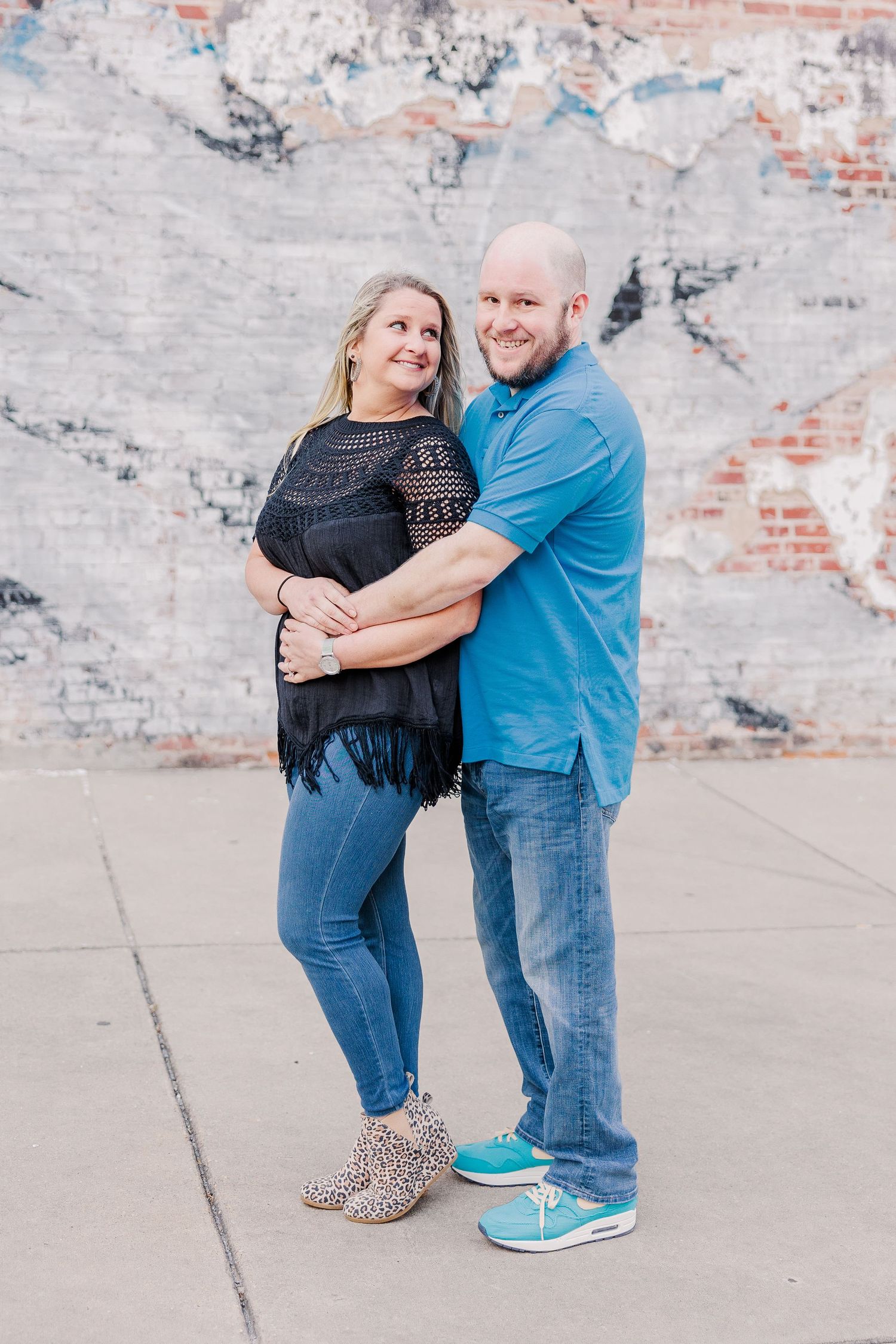 A couple hugs and smiles while posing against an industrial brick wall backdrop.