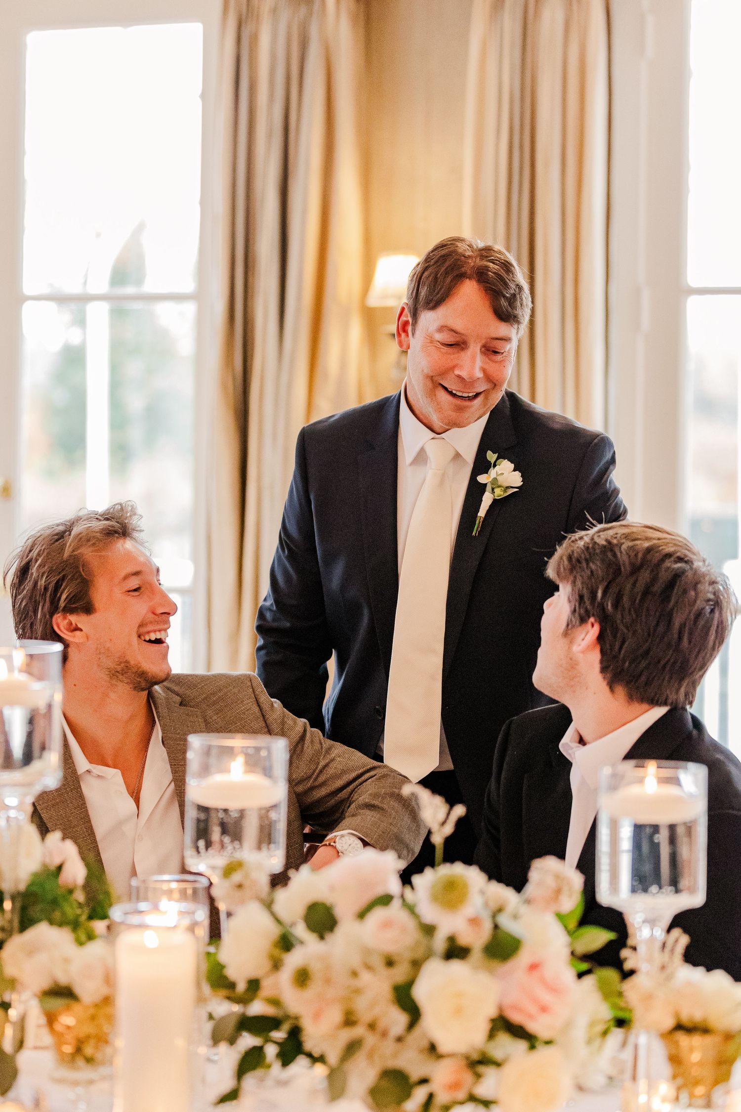 Three guests sharing cheerful moment at formal dinner with floral centerpieces.
