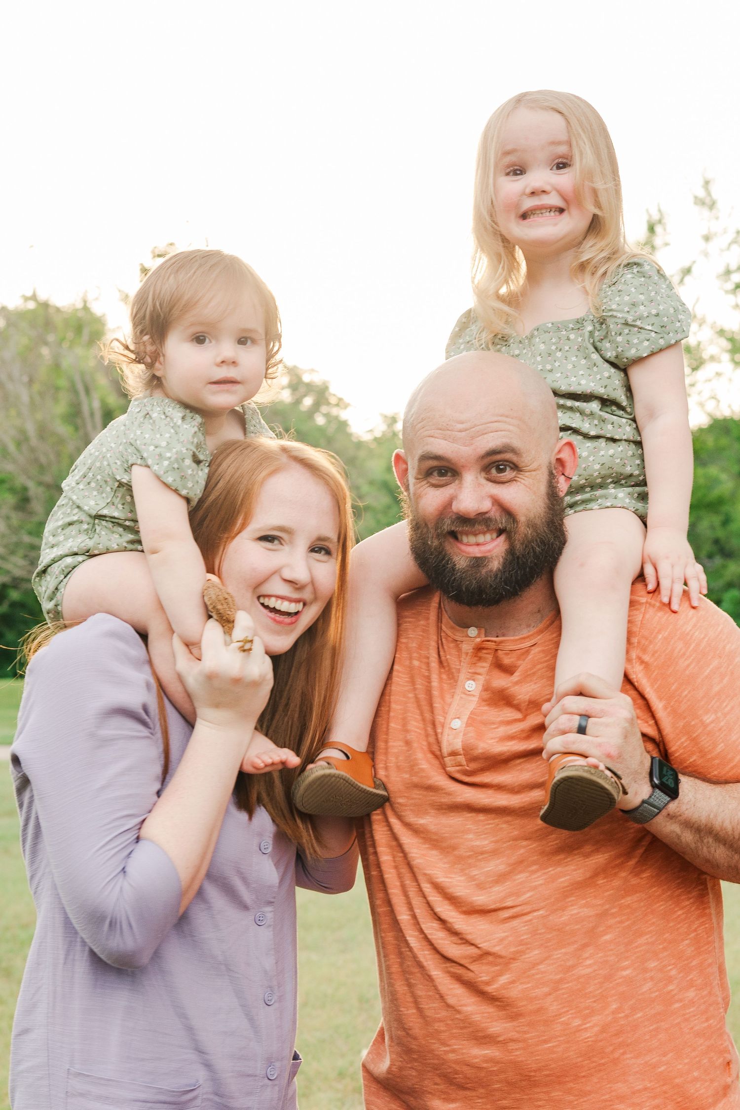 A happy family of four posing together outdoors with children on shoulders.