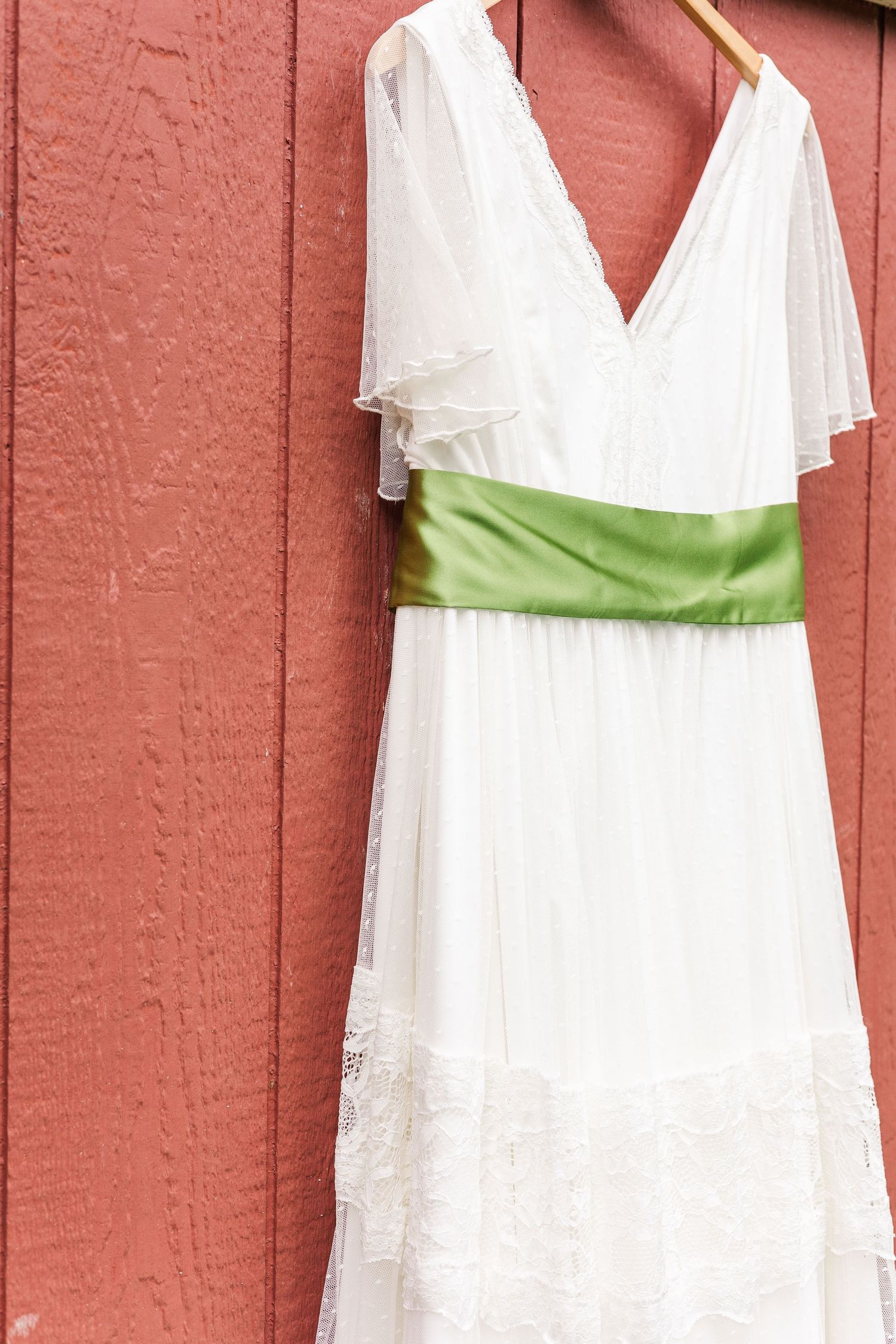 White wedding dress with flutter sleeves and green sash belt hangs against coral colored wall.