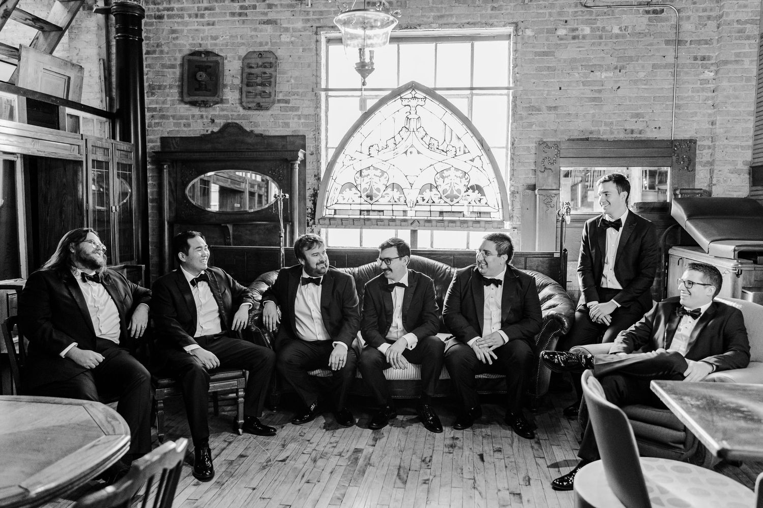 A black and white photo of groomsmen in tuxedos seated together in front of a decorative window.