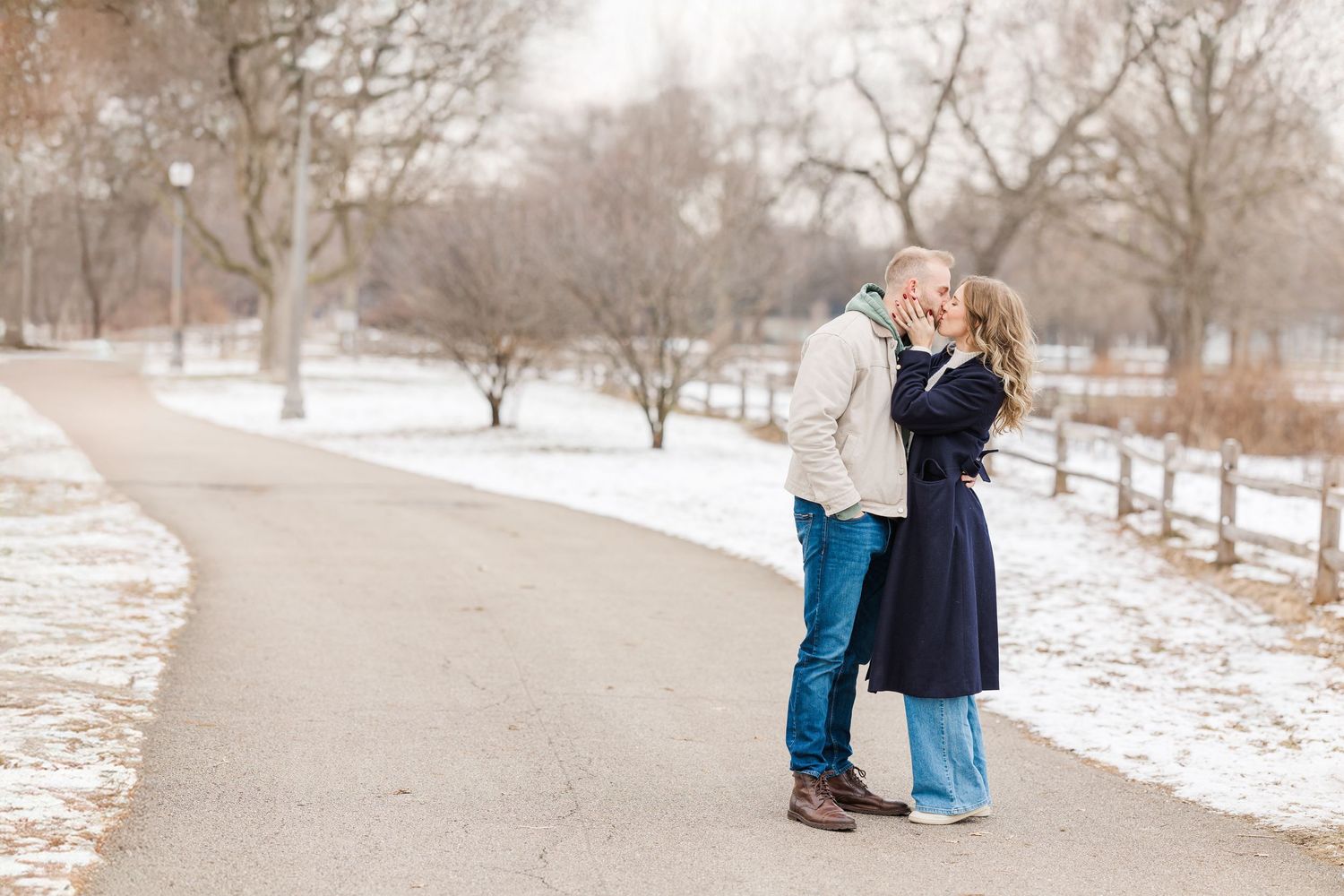 A couple shares a tender moment on a snowy winter path lined with bare trees and white fencing. winnemac Park Chicago
