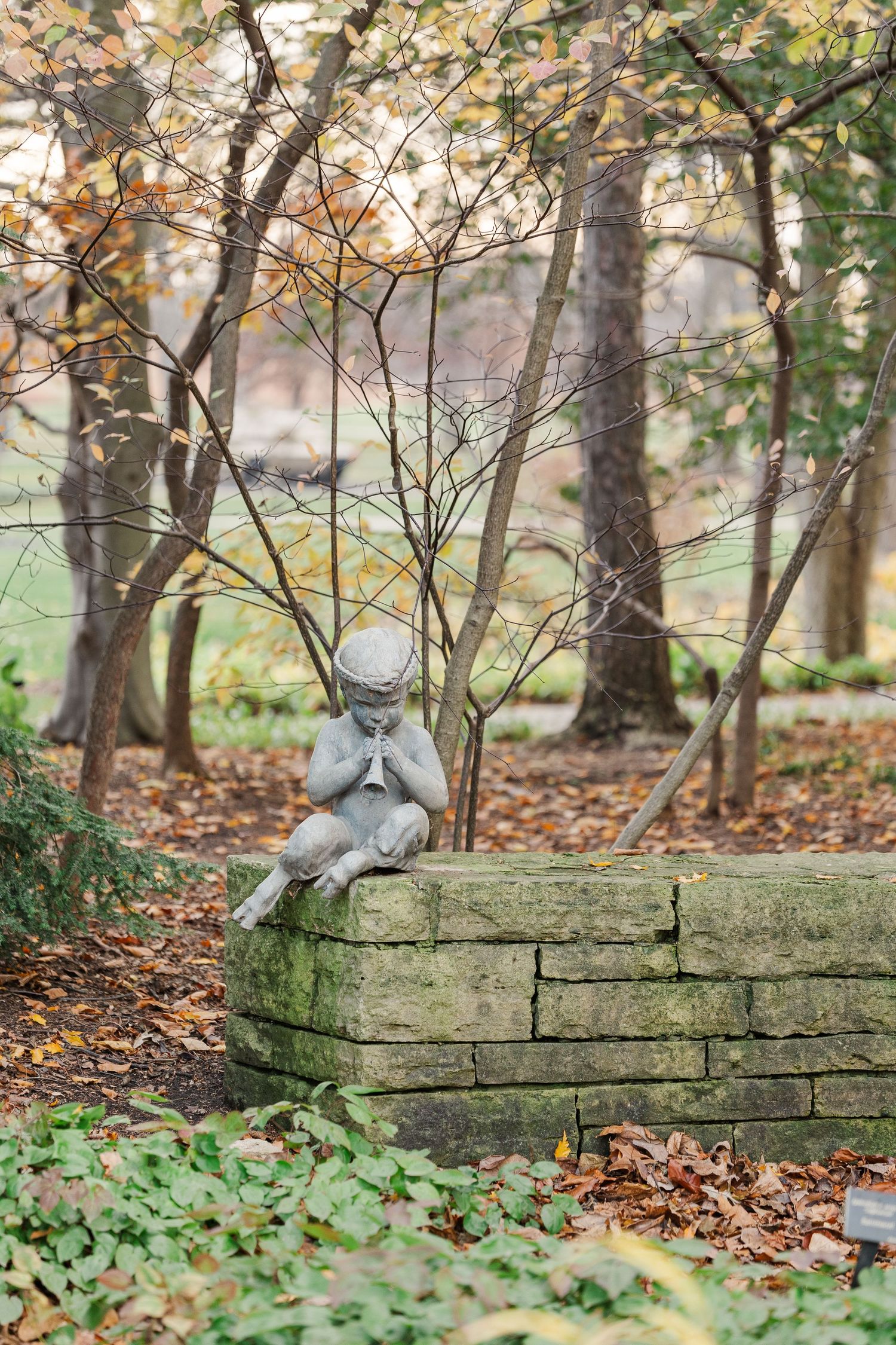 A stone cherub statue sits on a mossy garden wall surrounded by autumn leaves and bare trees in a peaceful setting.