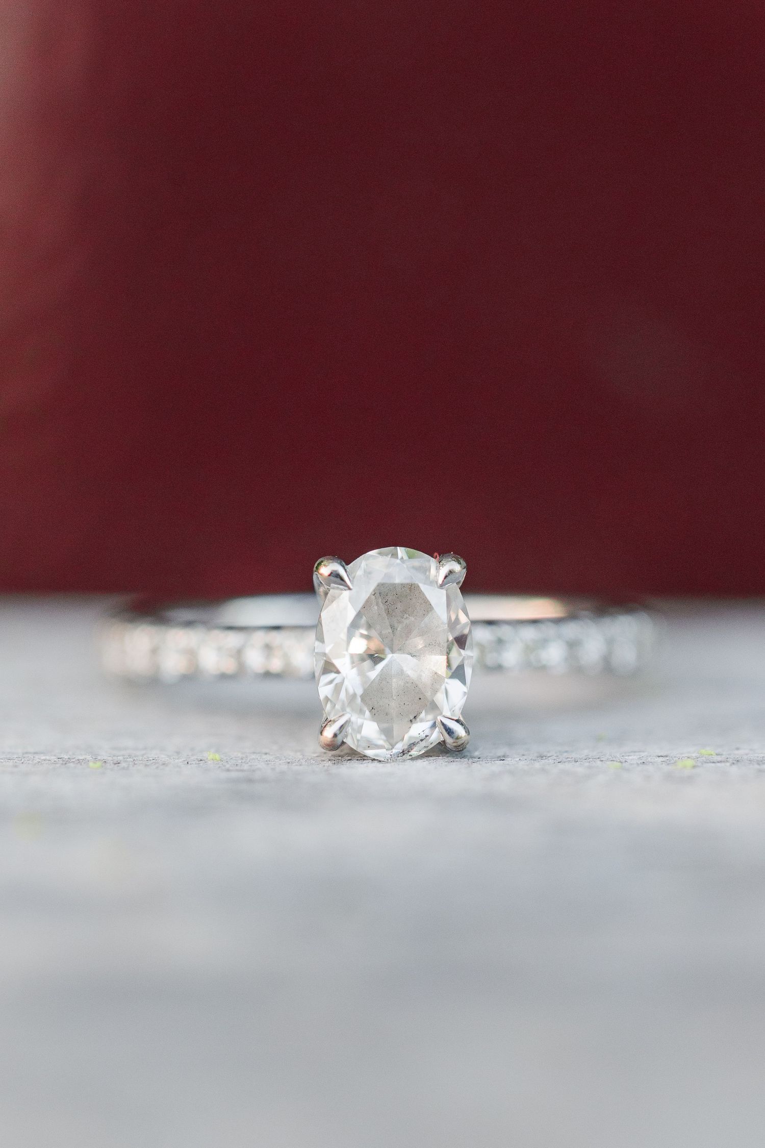 Close-up of an oval diamond engagement ring on a textured gray surface against a burgundy background.