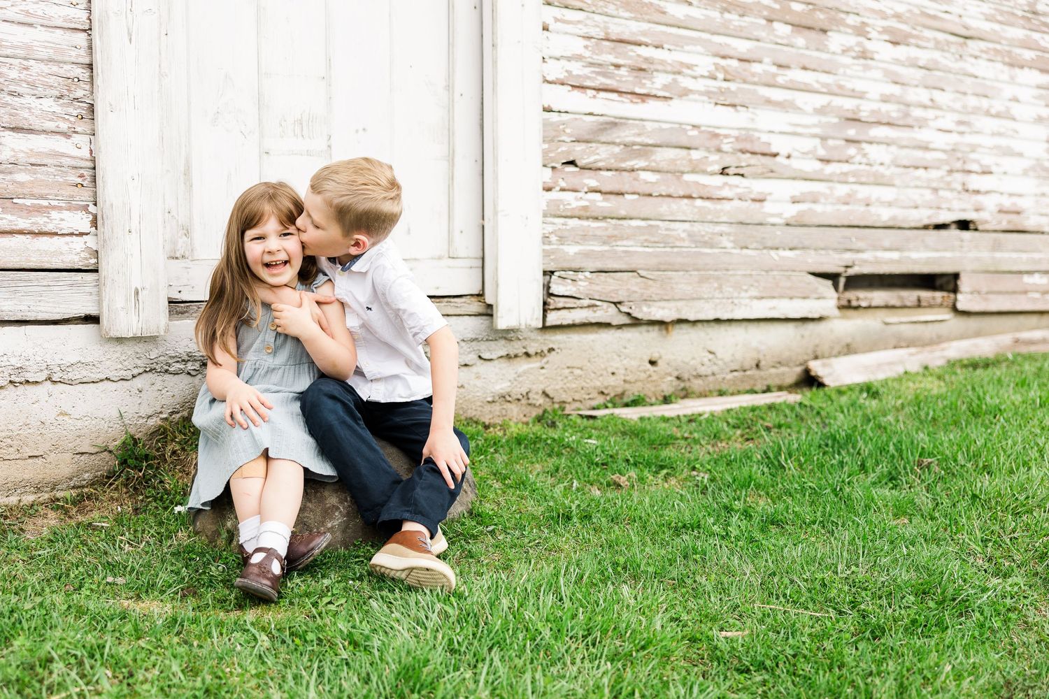 Two children share a playful moment while sitting together against a rustic white wooden wall.