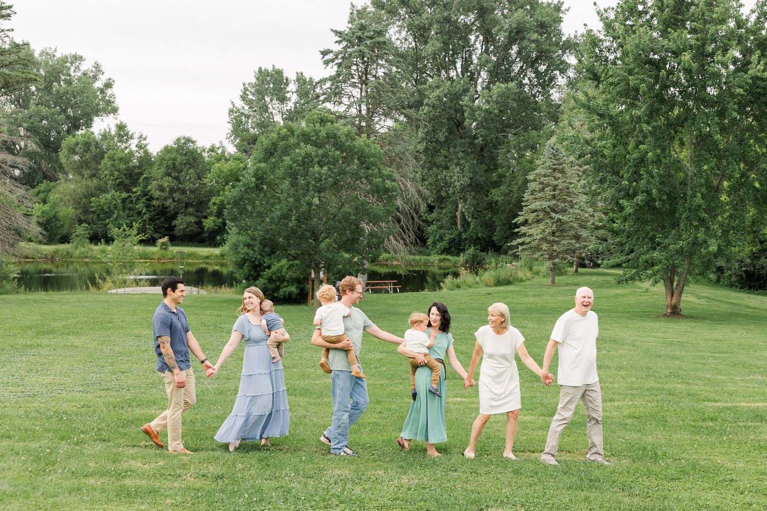 Extended family of seven walking hand-in-hand across a lush green lawn surrounded by trees.