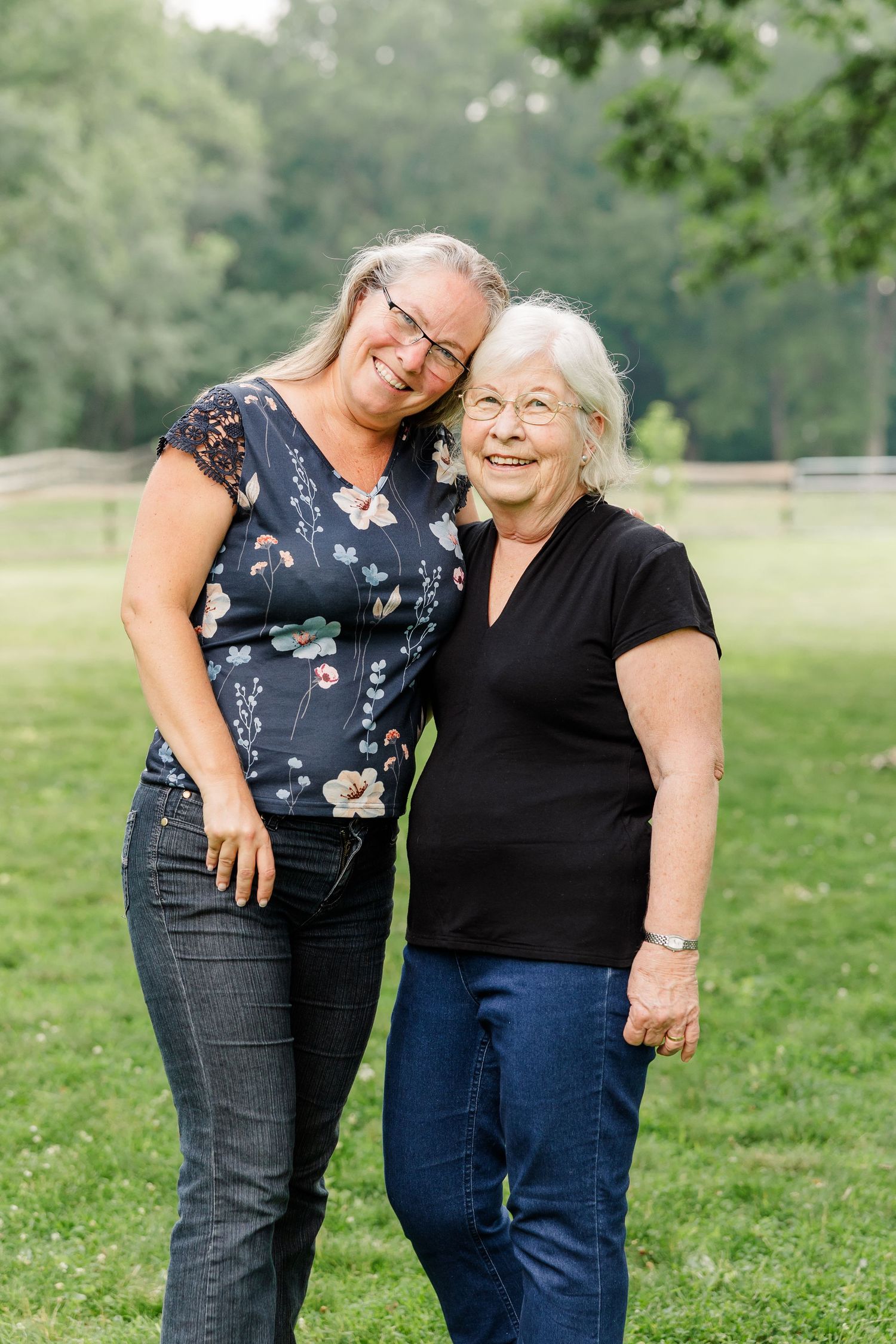 Two women smiling and embracing outdoors in a green park, wearing casual clothes.