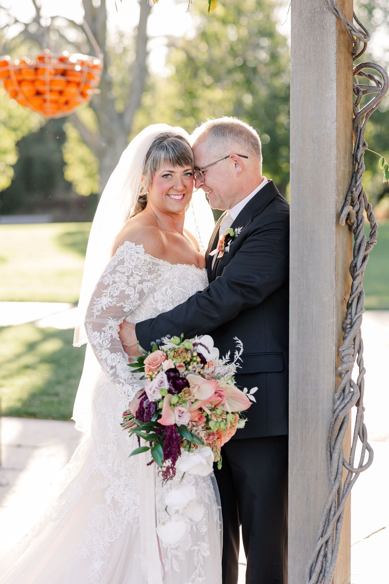 Smiling bride in a lace wedding gown and veil holds a colorful bouquet while embracing her groom in a dark suit outdoors.