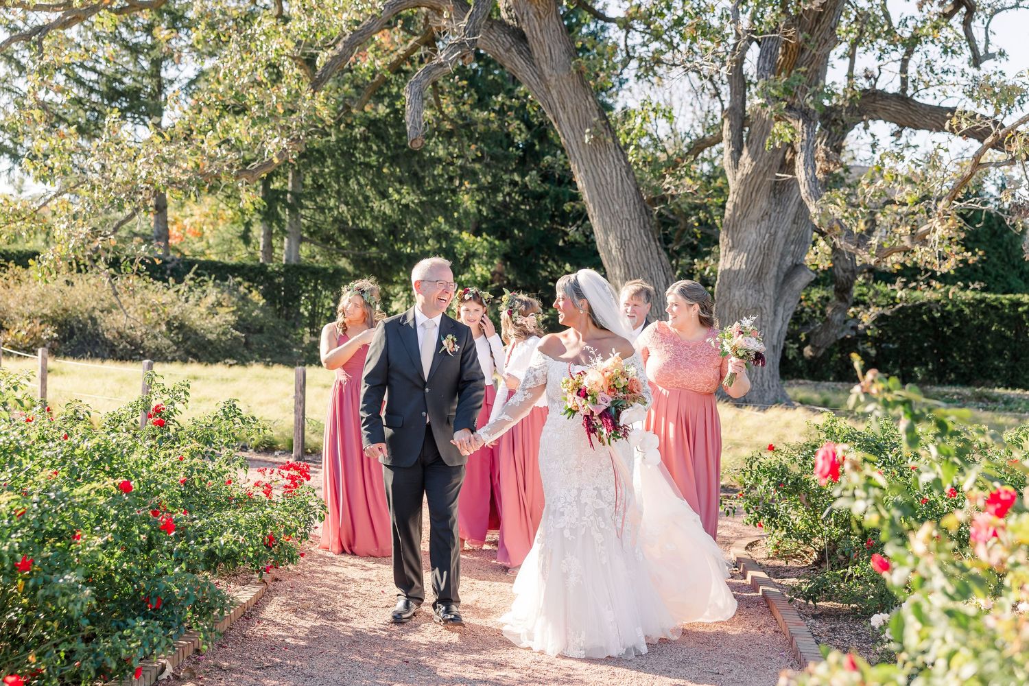 A groom in a dark suit walks hand in hand with his bride in a lace gown through a rose garden, followed by bridesmaids in pink dresses.