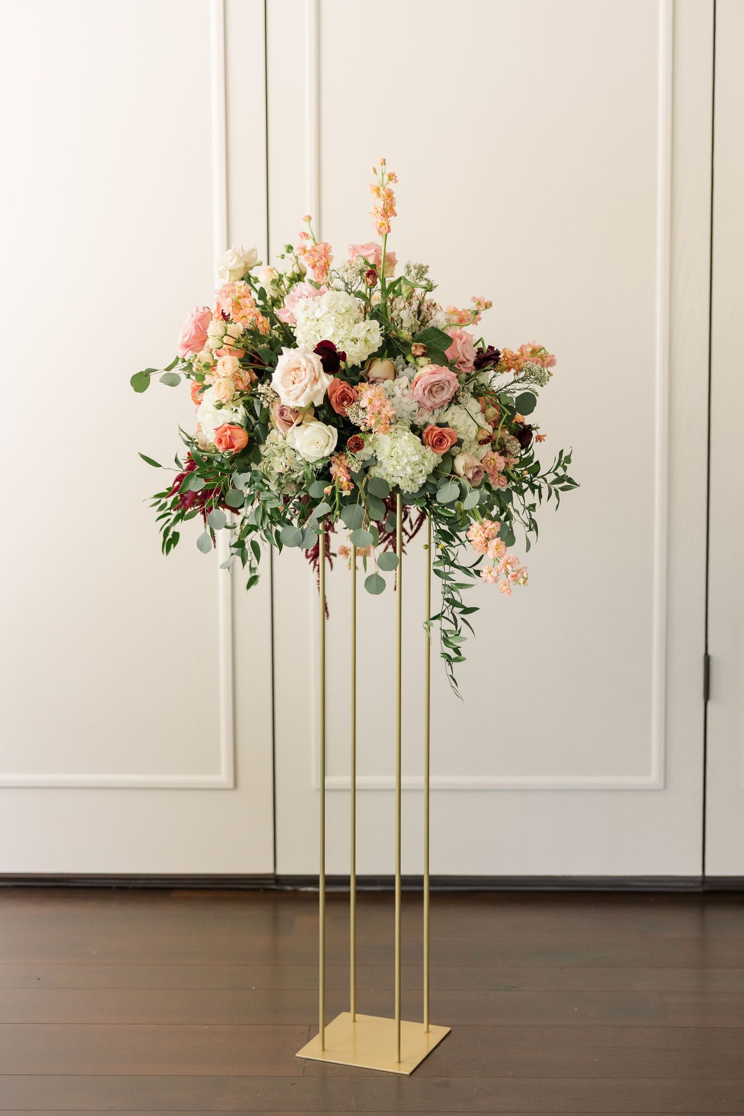 Tall gold stand wedding centerpiece with blush, peach, and white roses and hydrangeas with eucalyptus greenery on a wood floor.