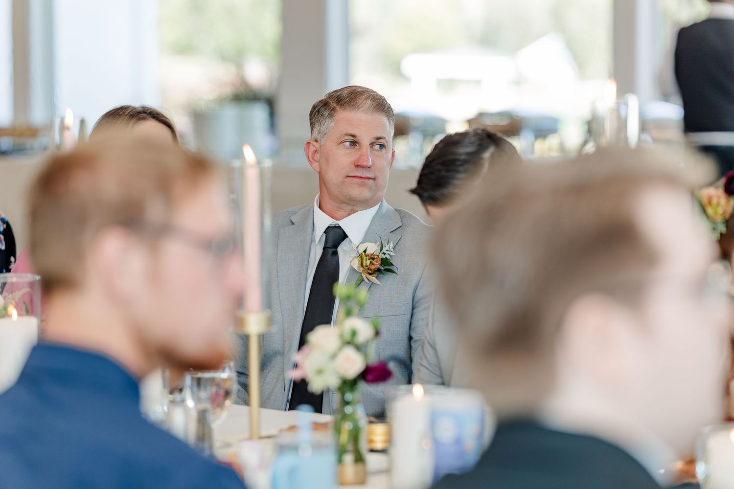 Man in a gray suit with a floral boutonniere sits at a wedding reception table with candles and flowers while guests surround him.