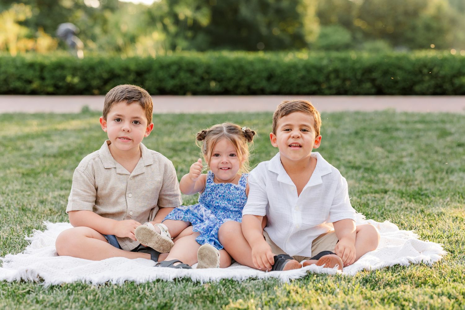 Three young children sitting on a white blanket on green grass in a sunny park, smiling for a family photo.