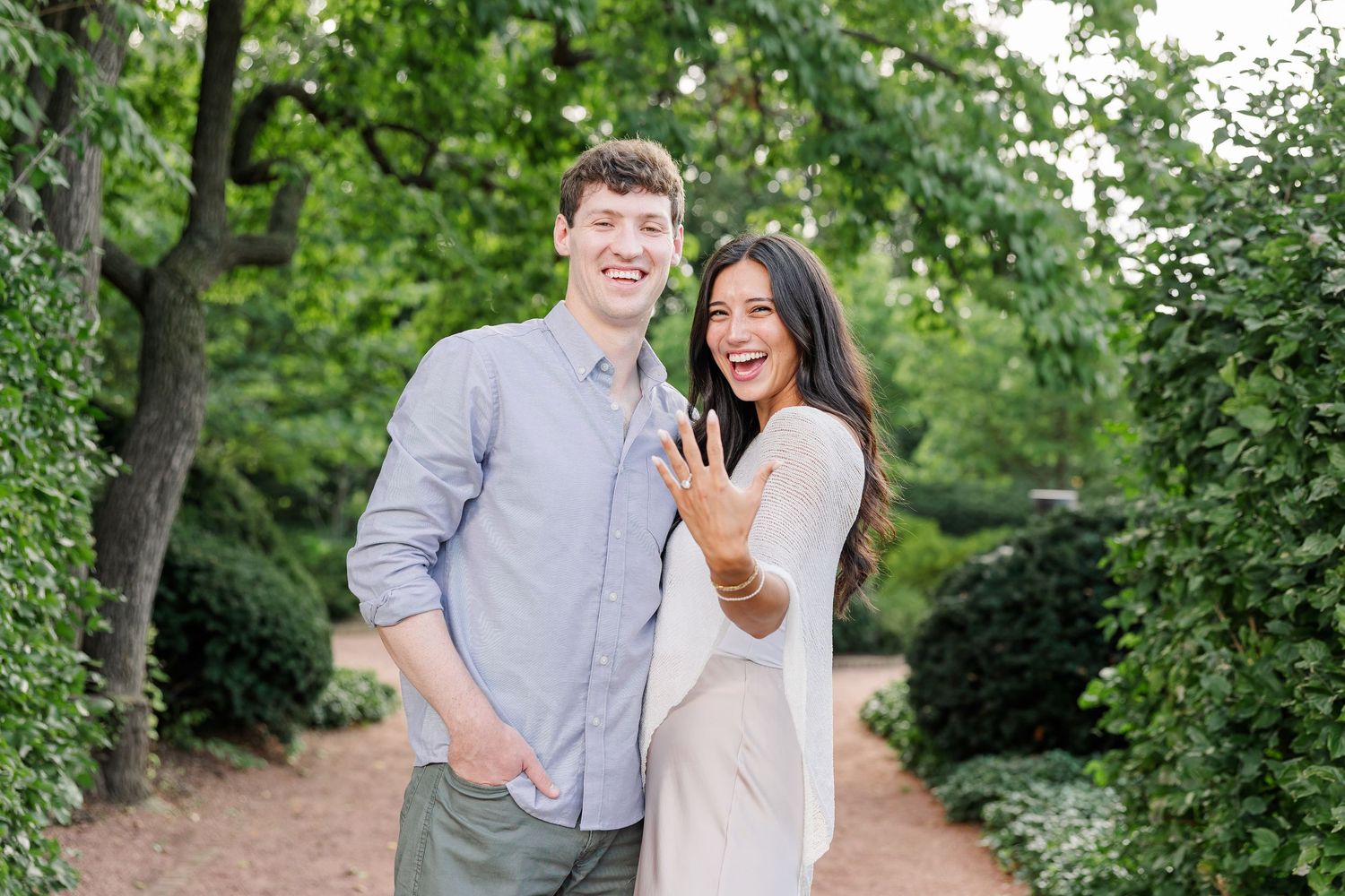 Happy couple posing outdoors showing off engagement ring, smiling in lush green garden setting.