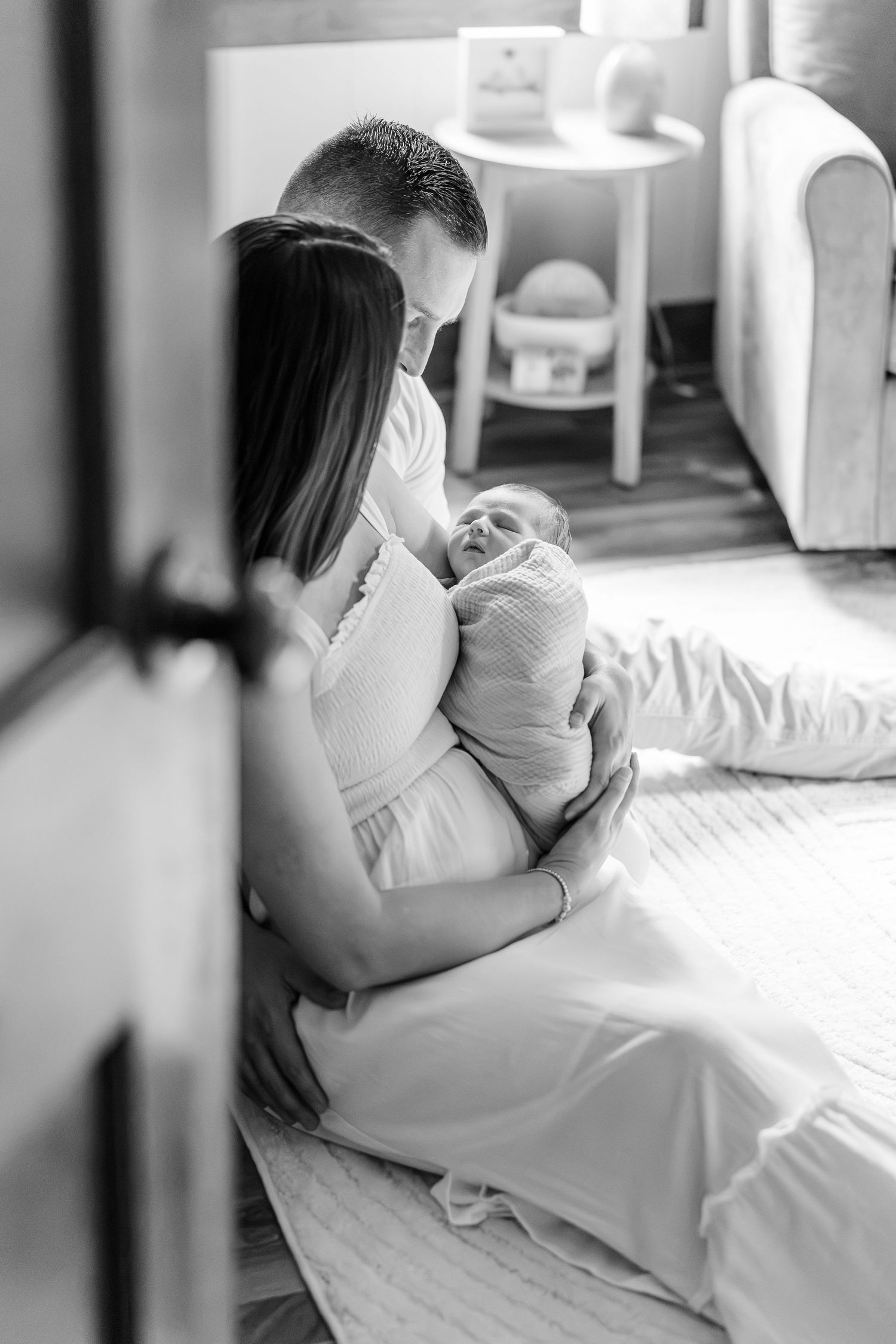 Black and white photo of parents sitting on floor together, lovingly cradling their newborn baby at home.