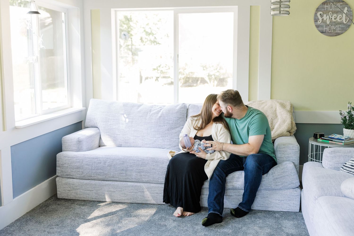 A couple sits on a light gray sofa, tenderly gazing at their newborn baby in a bright, cozy living room.