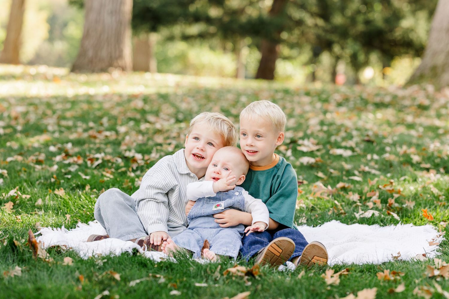 Three young siblings sit together on a blanket in a park during autumn, smiling joyfully outdoors.