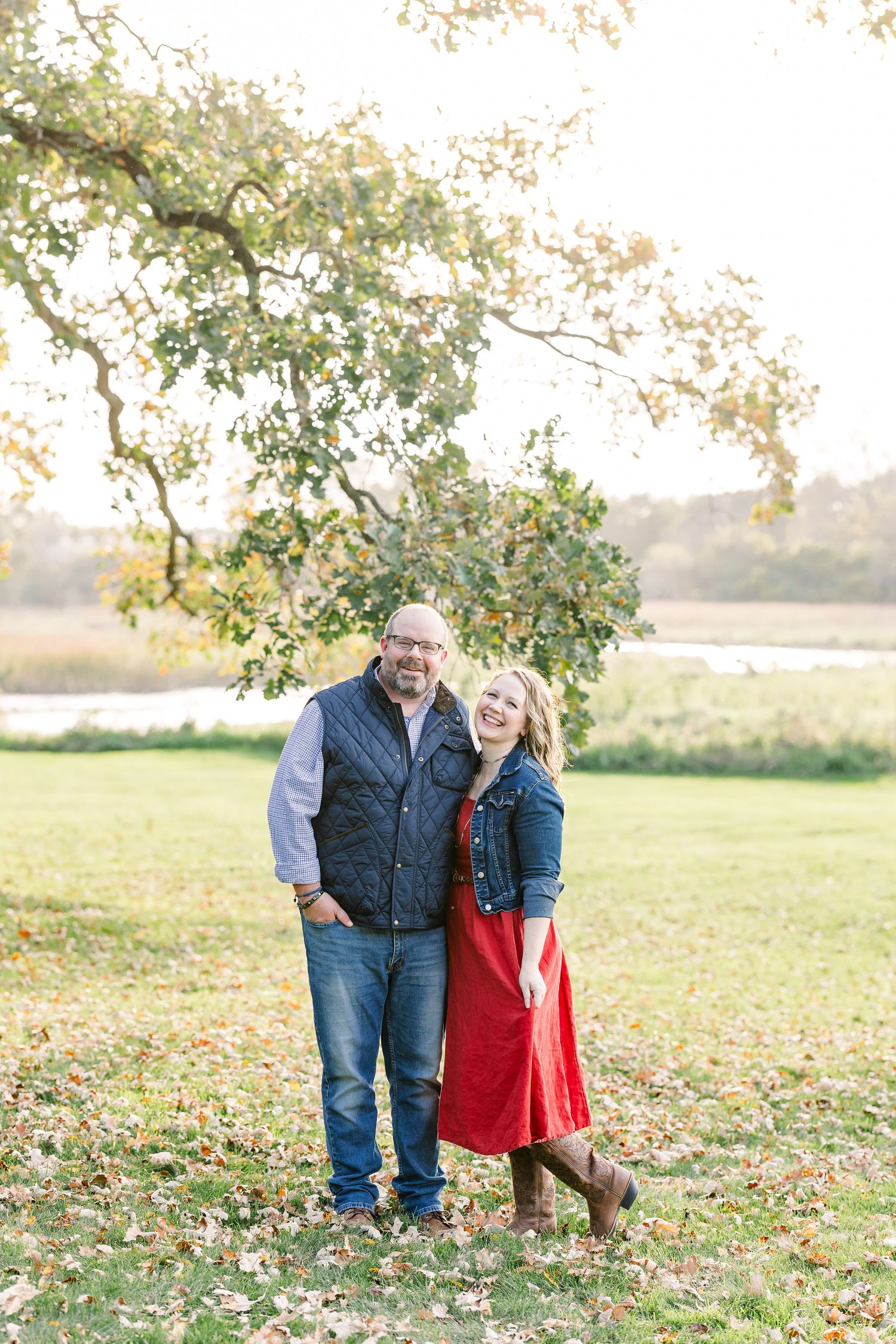 A smiling couple in casual fall outfits stands arm in arm on a grassy lawn beneath a tree near a pond during family portraits.