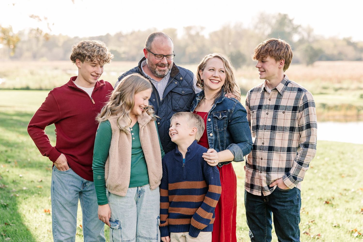 Smiling family of six, a man, a woman and four children, posing together and laughing in a grassy field during an outdoor portrait session.