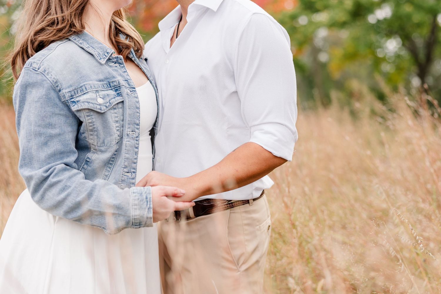 Couple holds hands in a tall golden grass field, woman in denim jacket and white skirt beside man in white shirt.