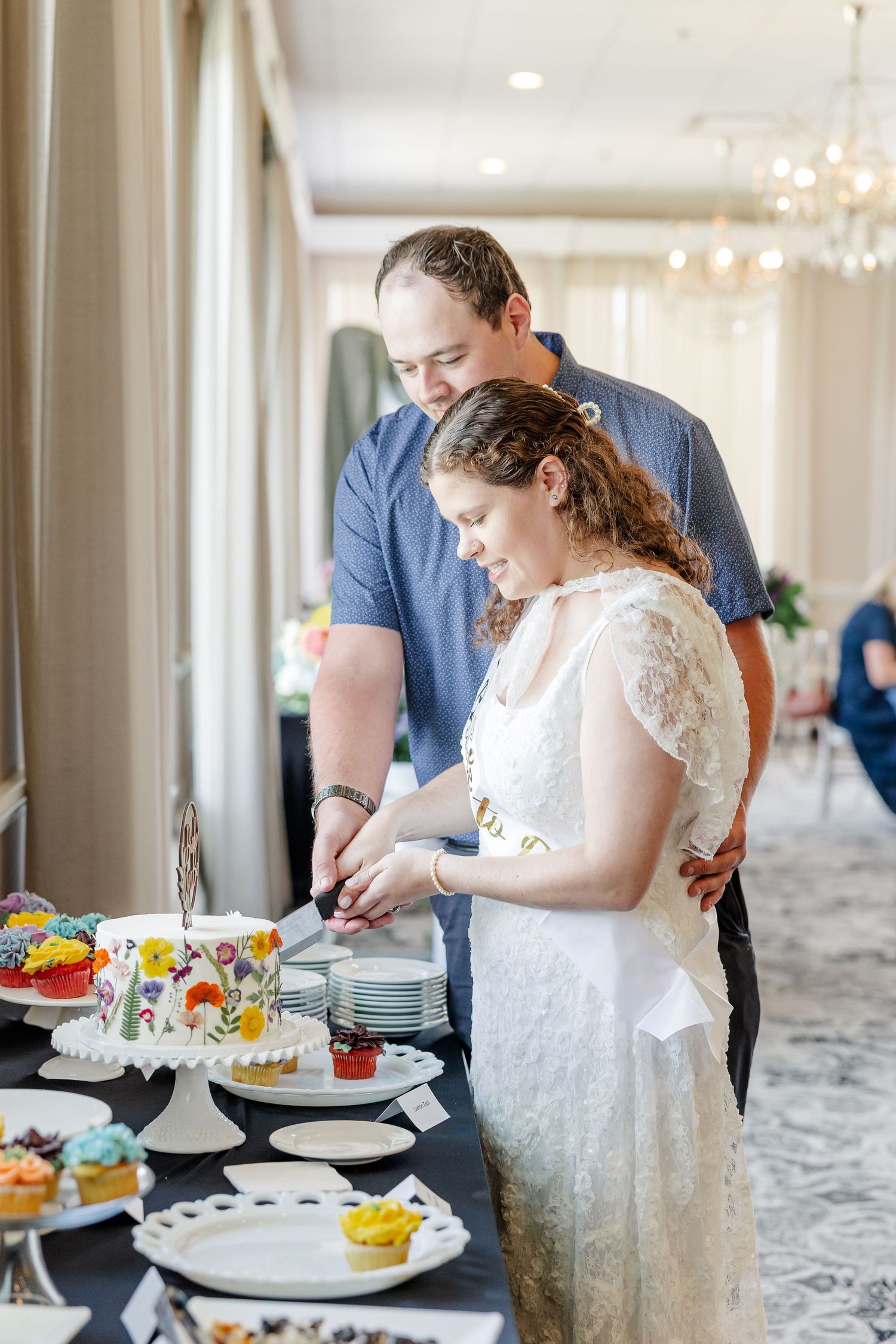 Bride and groom cutting a floral decorated wedding cake at a dessert table with colorful cupcakes.