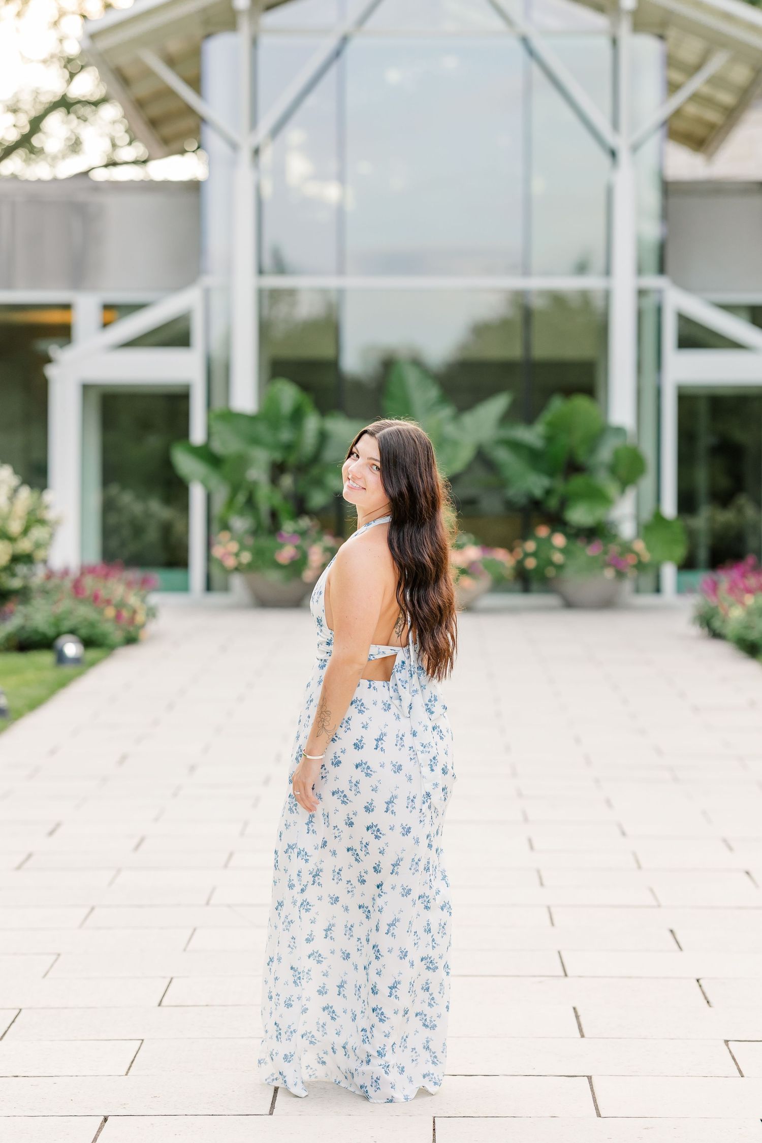A young woman in a floral maxi dress smiles over her shoulder in front of a modern glass building with lush greenery.