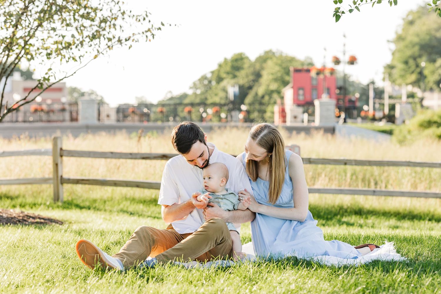 A young family sits on grass outdoors, parents lovingly attending to their baby on a sunny day near a wooden fence.