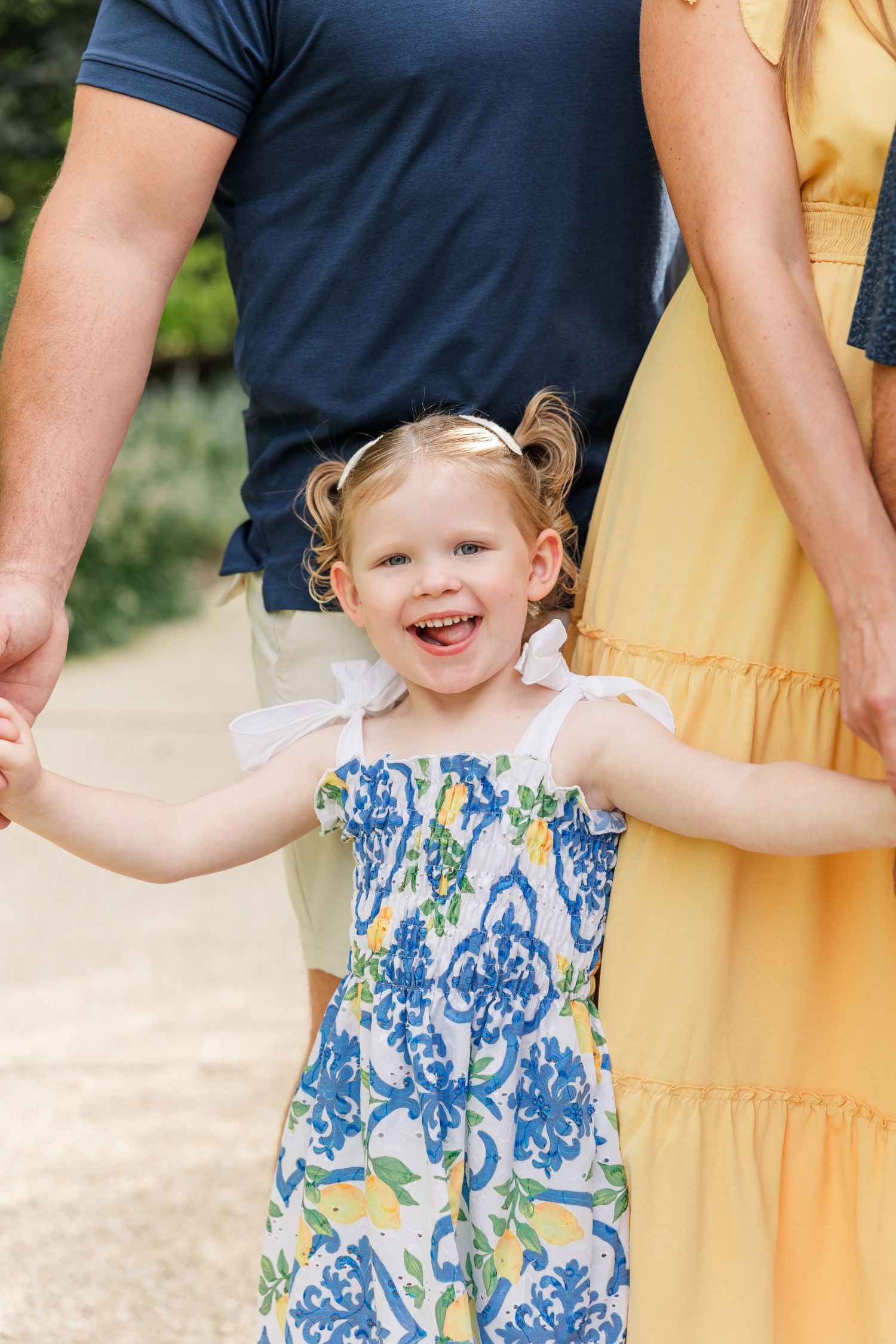 Laughing toddler girl in blue floral dress holds parents hands outdoors.