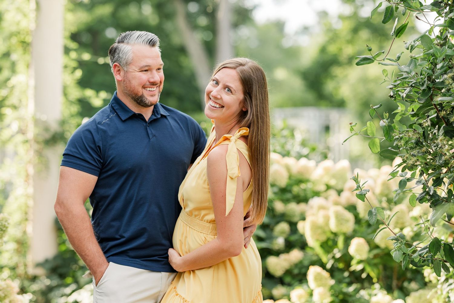 Smiling couple standing together near hydrangeas, woman in yellow dress with baby bump.