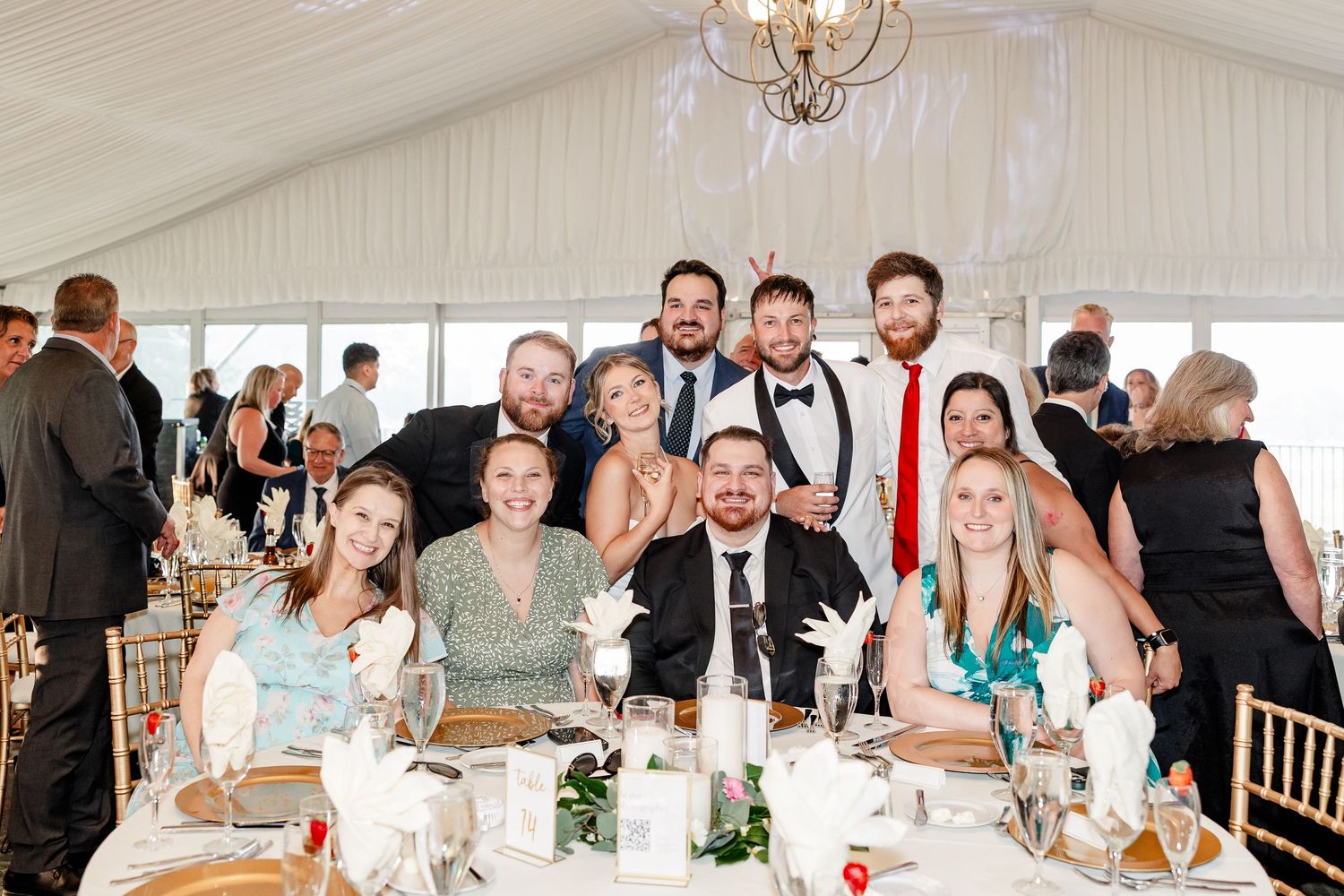 Large group of wedding guests smile together at a round reception table in a decorated tent venue.