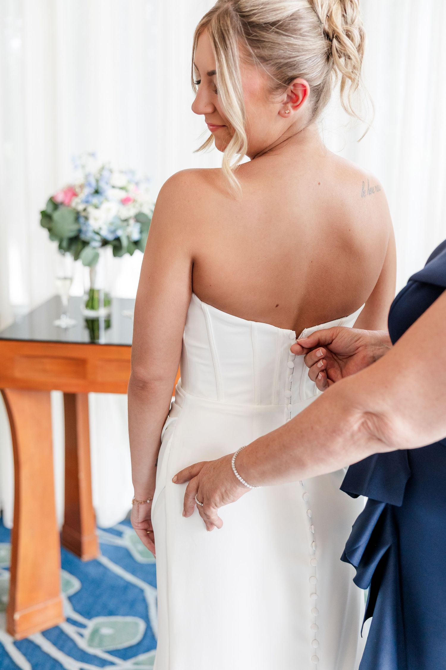 Bridesmaid buttoning the back of bride's strapless white wedding gown in elegant hotel room.
