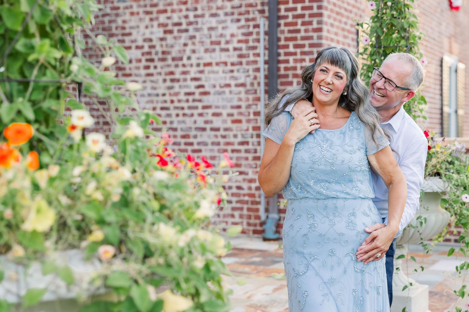Happy couple embracing outdoors near brick wall with colorful garden flowers blooming in background.