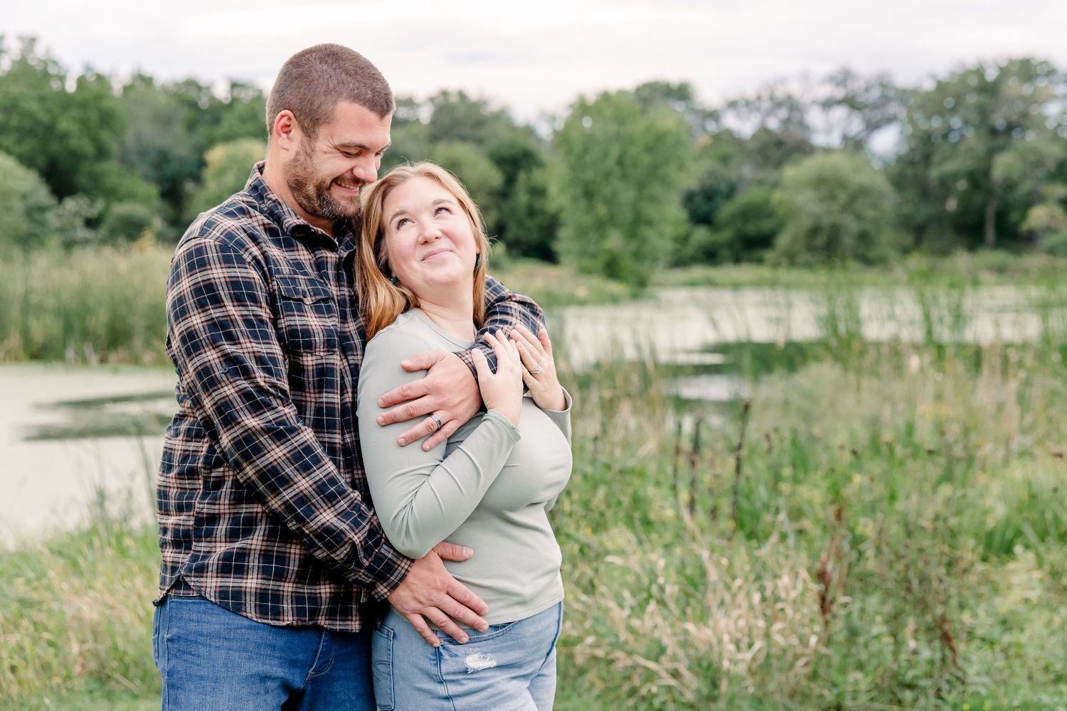 Happy couple embracing outdoors near a pond surrounded by lush green trees and tall grass.