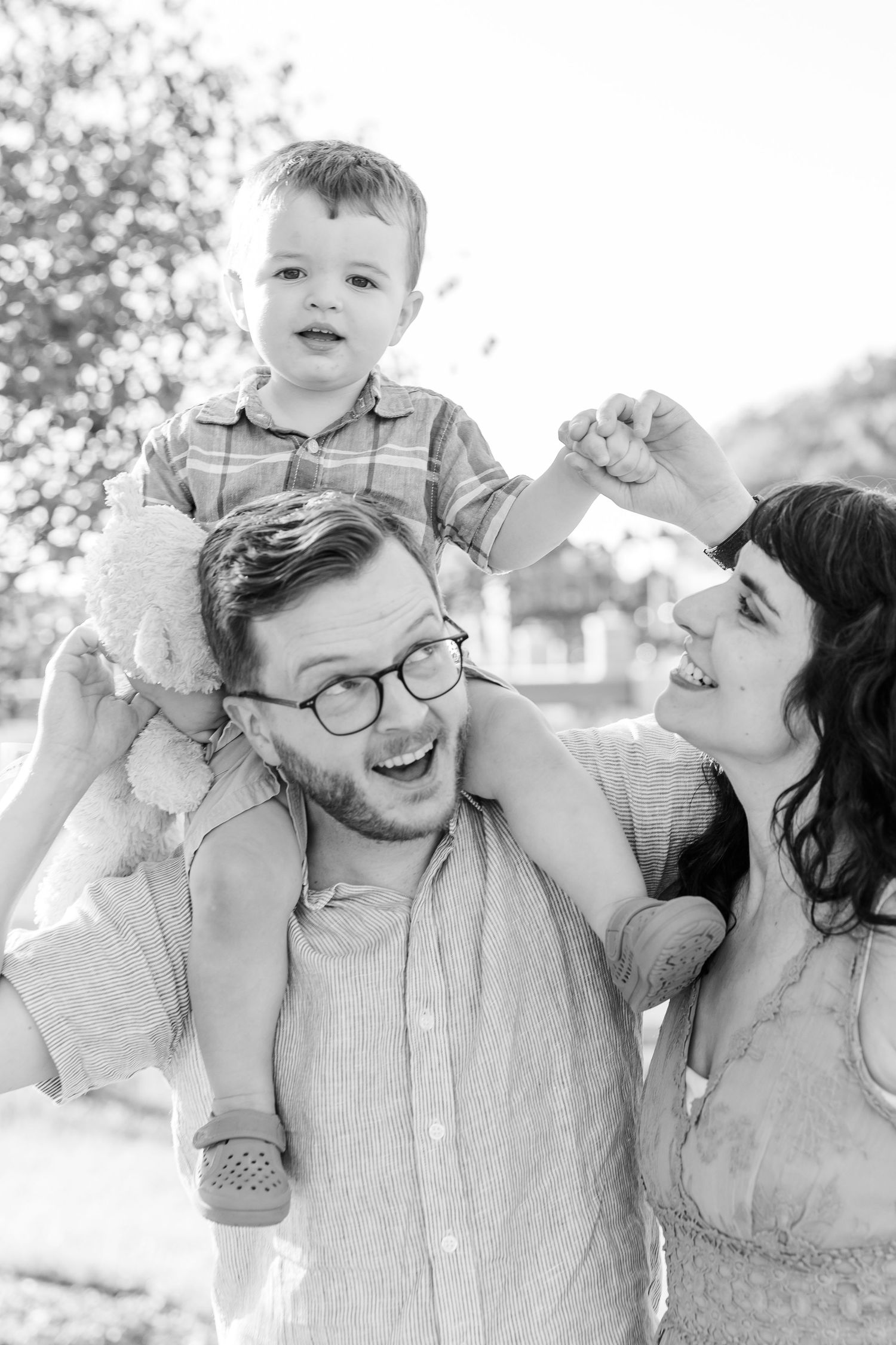 Black and white photo of happy family with dad carrying toddler on shoulders while mom smiles outdoors.