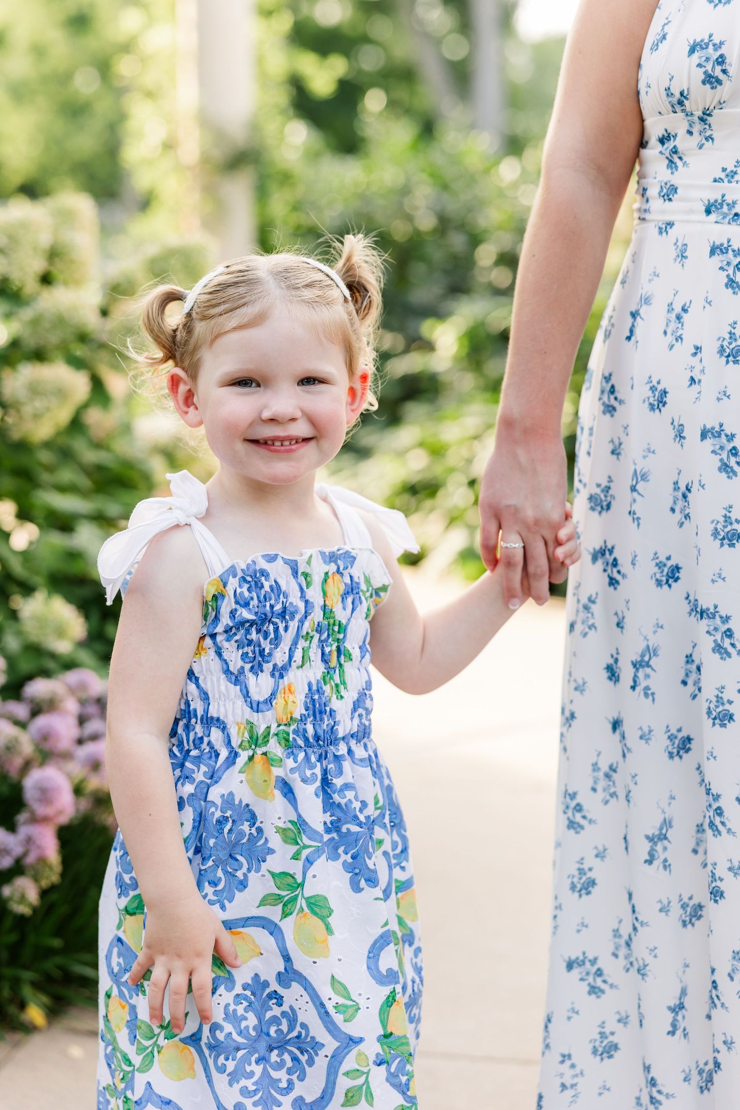 Smiling toddler girl in blue floral dress holds mothers hand in garden.
