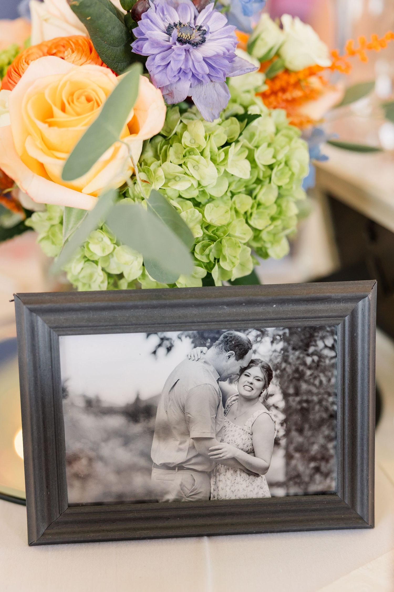 Black and white engagement photo in a gray frame beside a colorful floral wedding centerpiece.