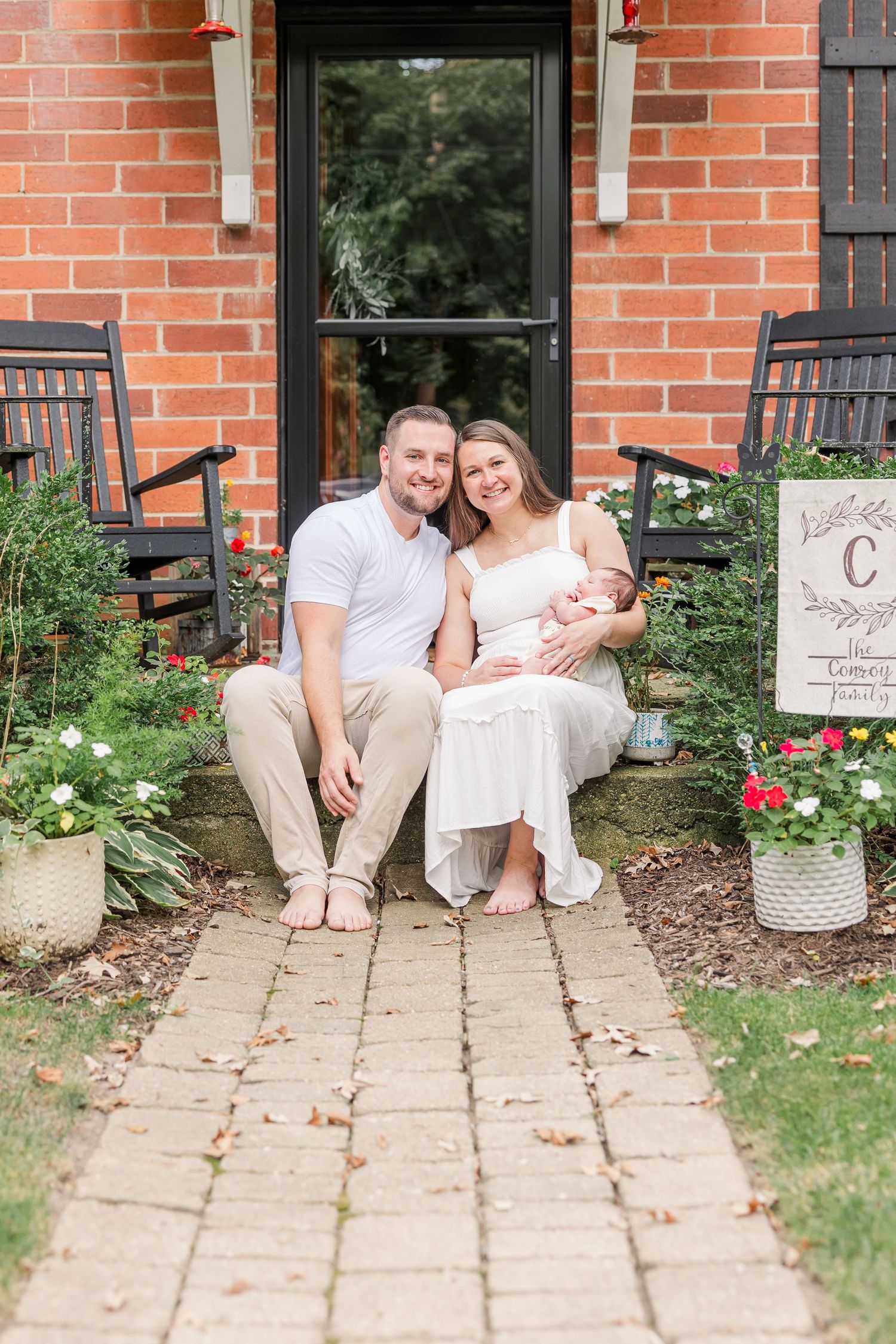 Smiling new parents sitting on brick front porch steps holding their newborn baby surrounded by colorful flowers.