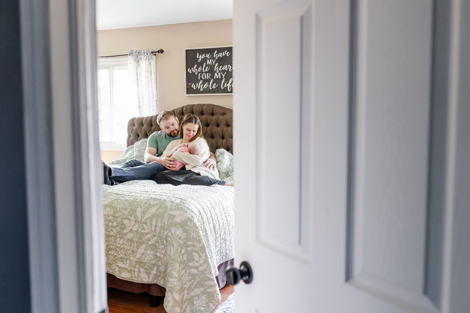 Couple sitting on bed together looking at newborn baby through open bedroom door in cozy home.