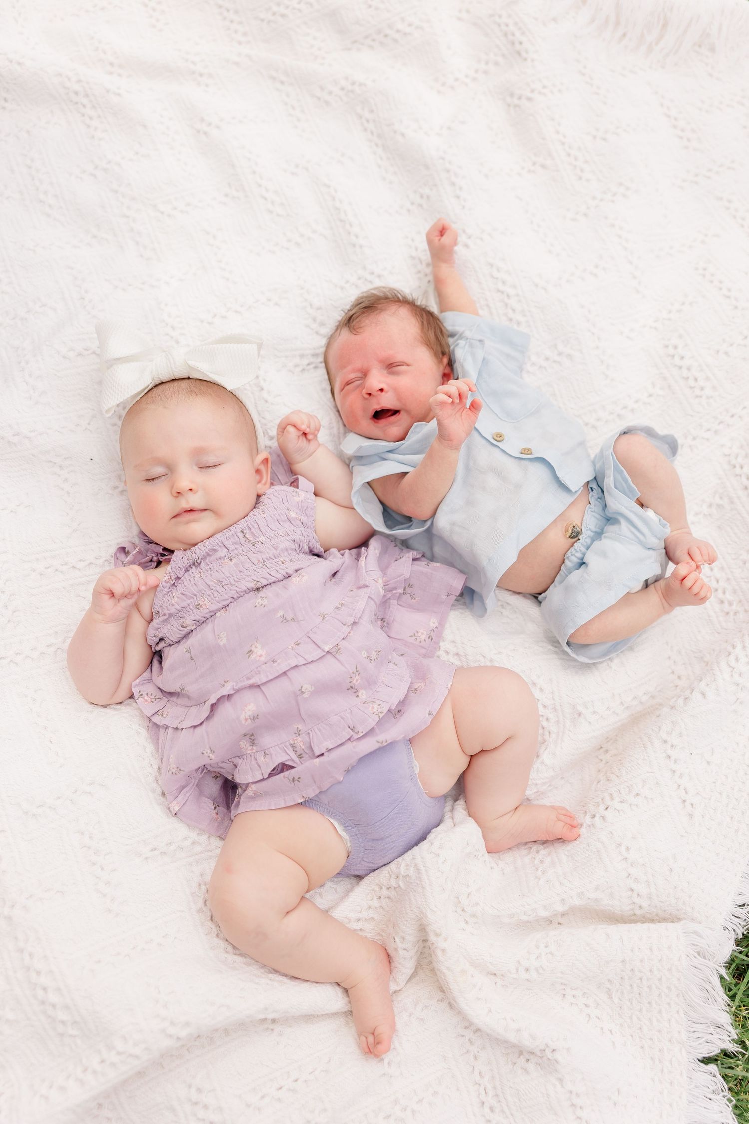 Two newborn babies lying on white blanket, one in purple outfit and one in blue, one crying.