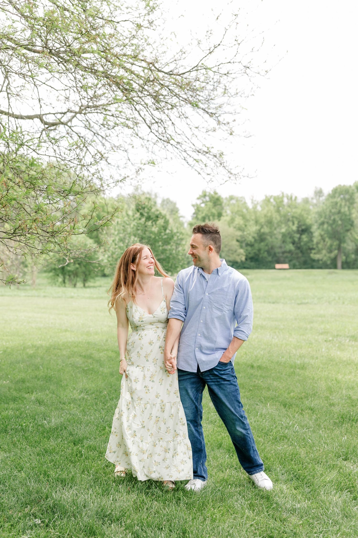 A happy couple holds hands and smiles at each other while walking in a lush green park in spring.