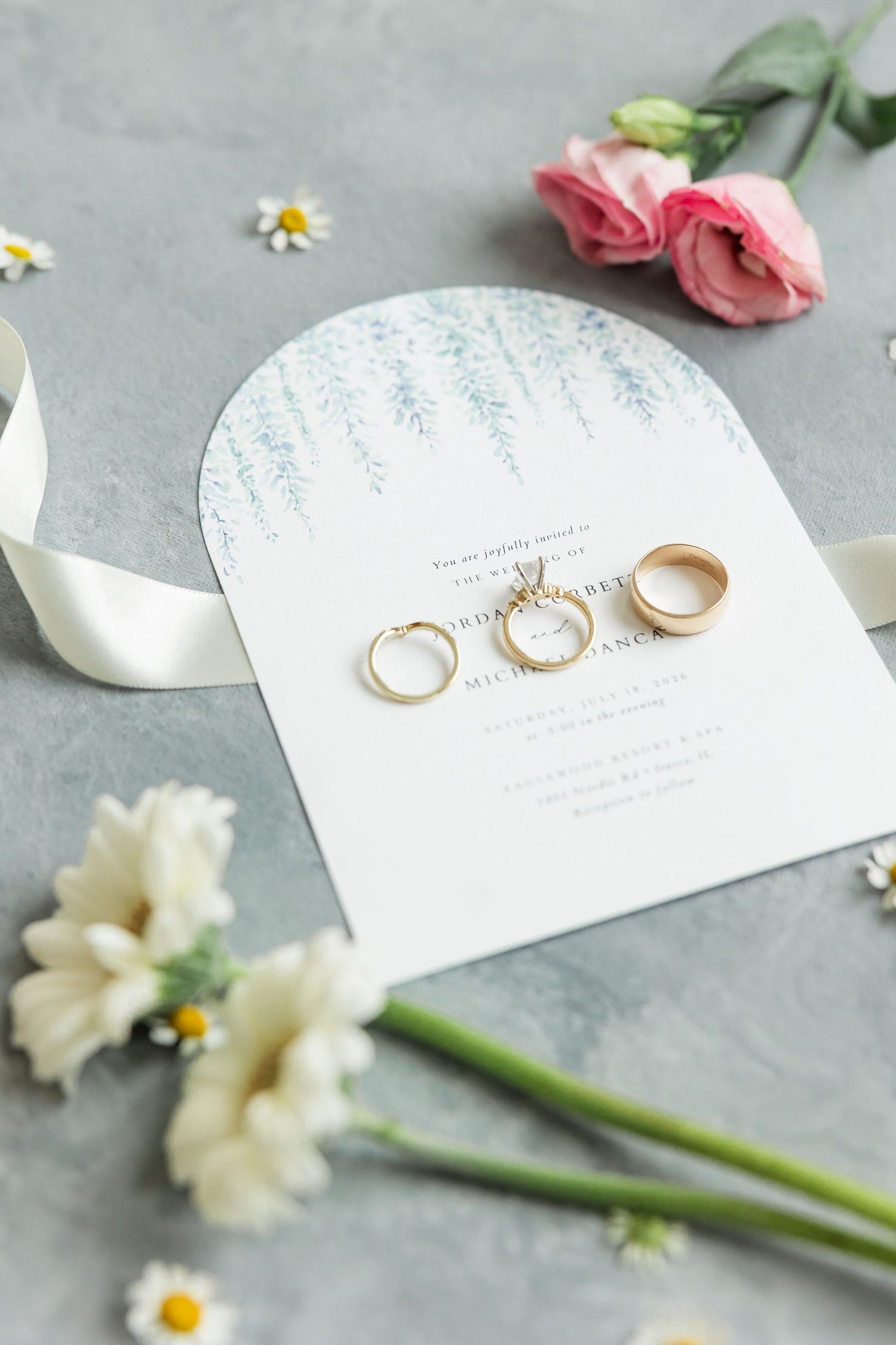 Gold wedding rings rest on an elegant arch-shaped wedding invitation surrounded by pink roses and white daisies on a gray surface.