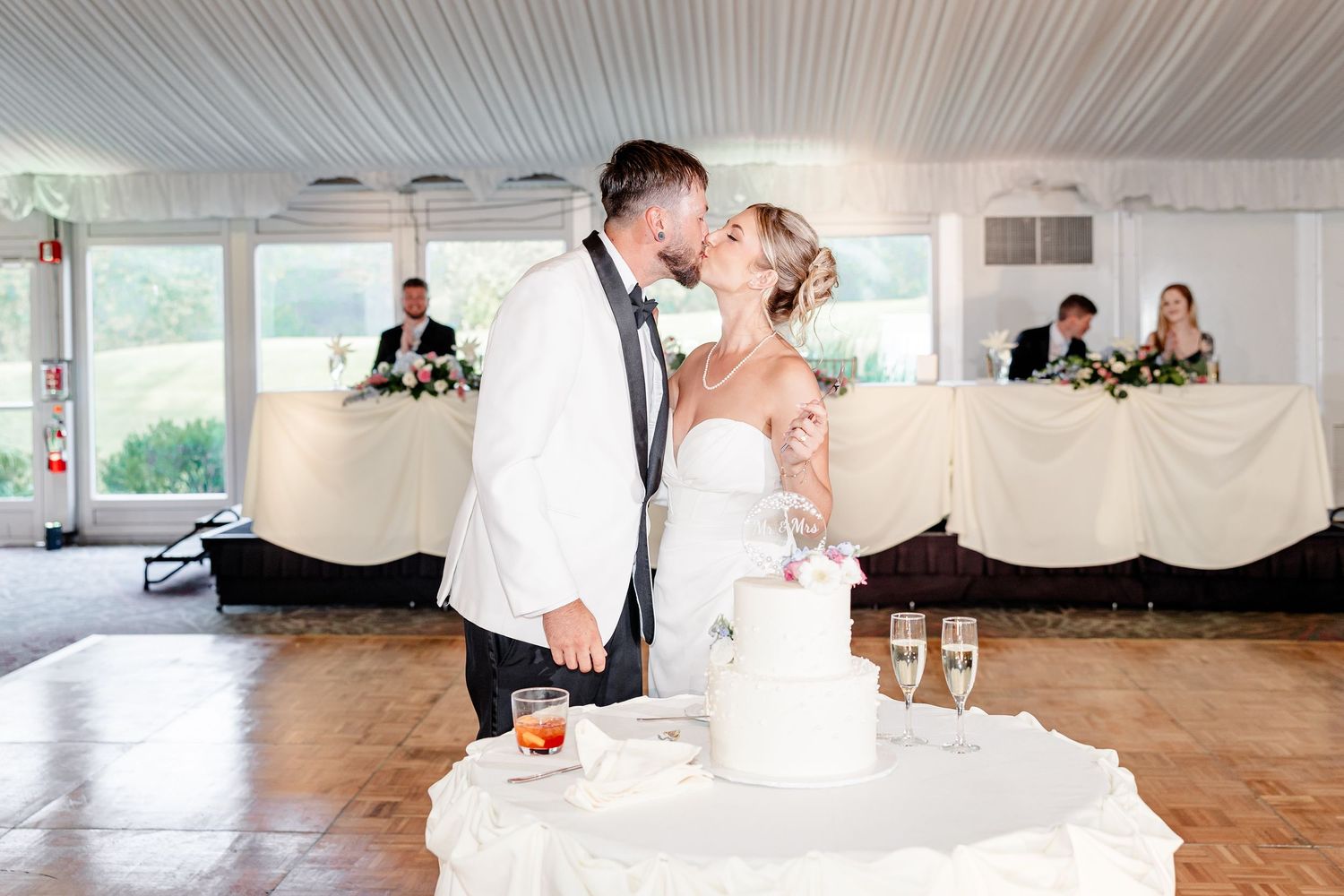 Bride and groom kiss beside white wedding cake at reception with head table in background.