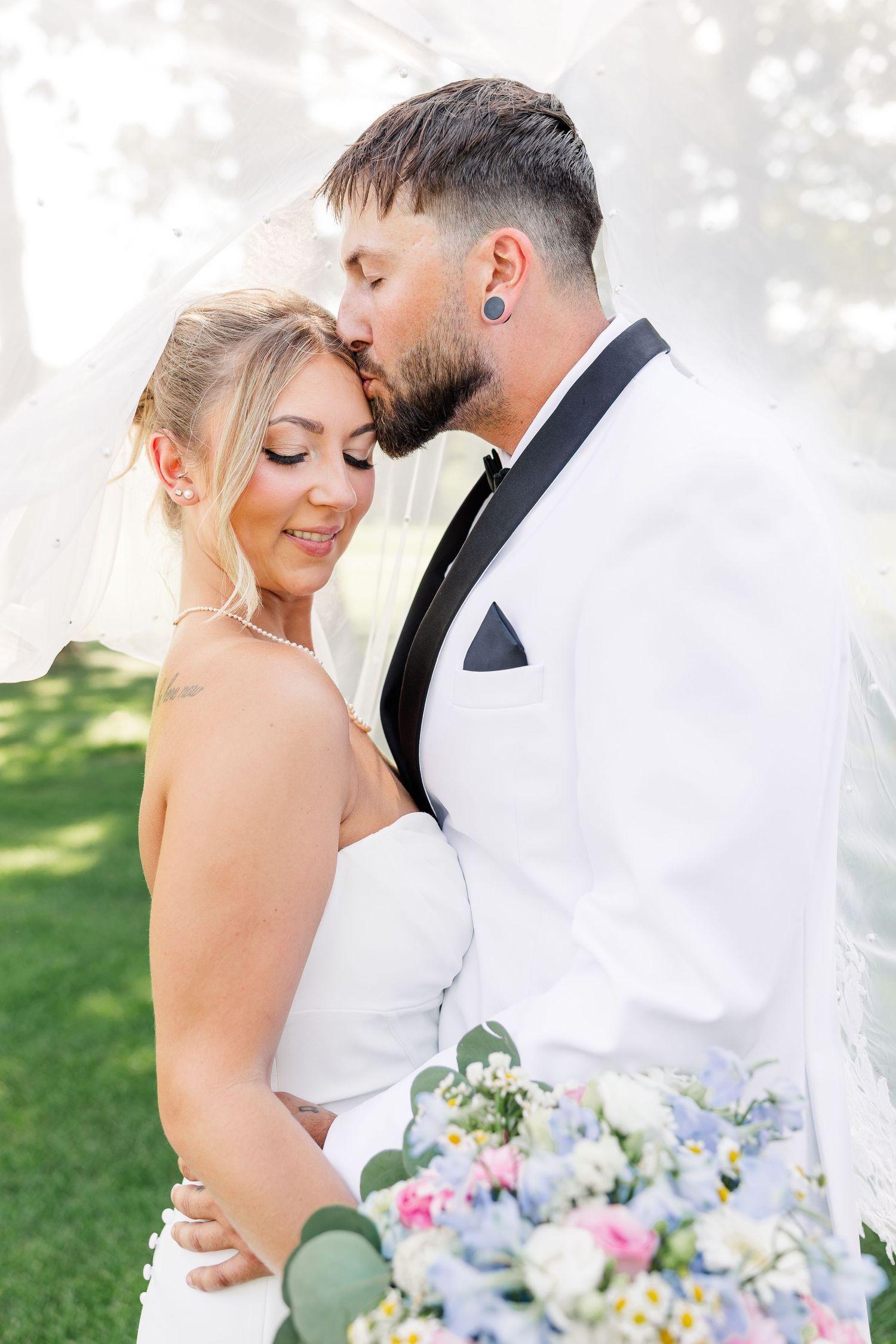 Groom in white tuxedo kissing bride on forehead while she holds a pastel floral bouquet.