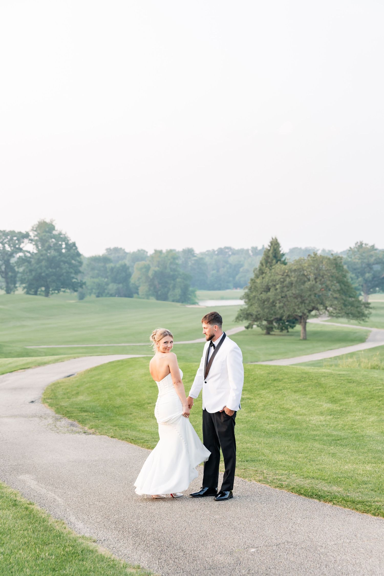 Bride and groom standing on a golf course path, bride in fitted white gown, groom in white tuxedo.