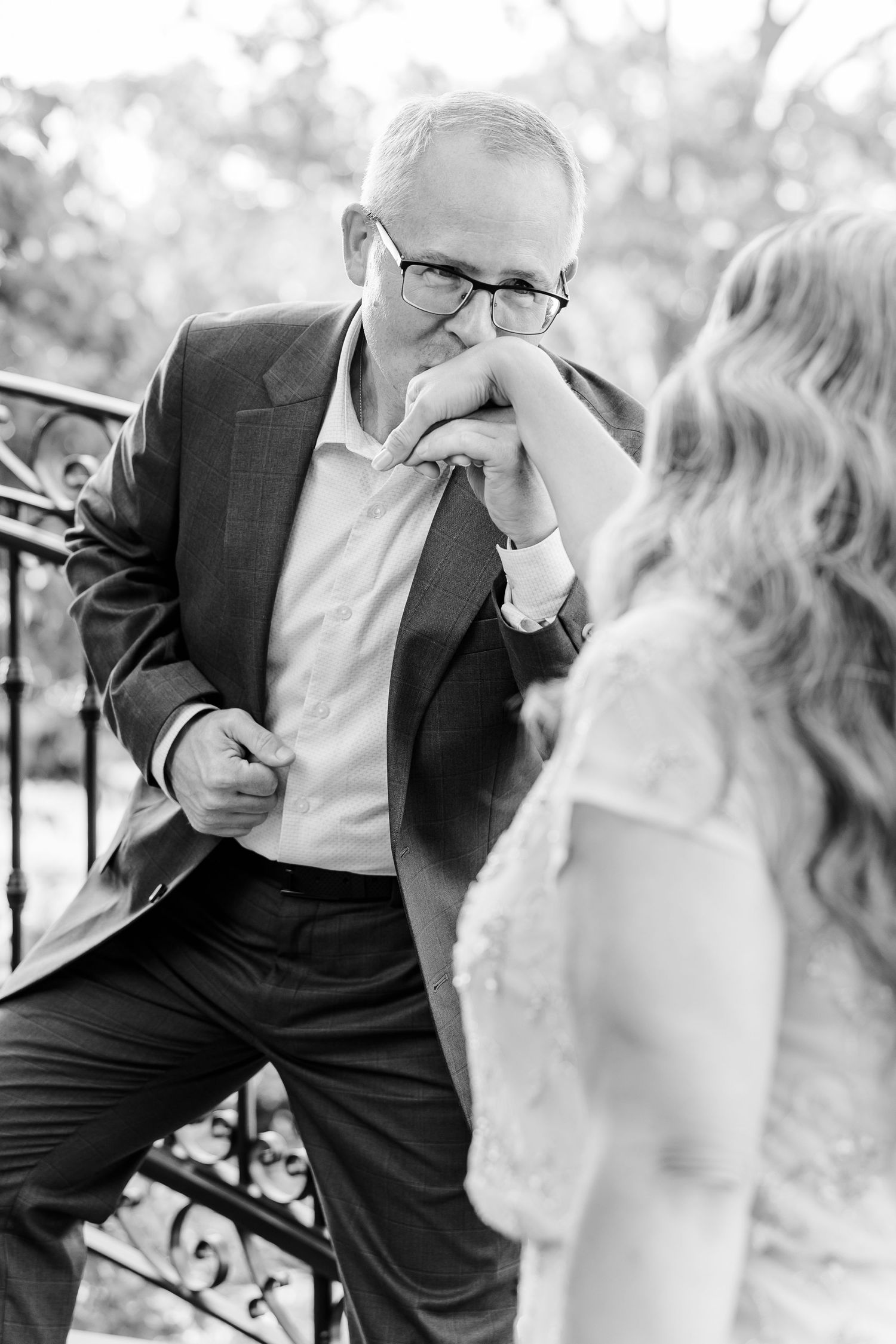 Black and white photo of a father kissing bride's hand on staircase at wedding.