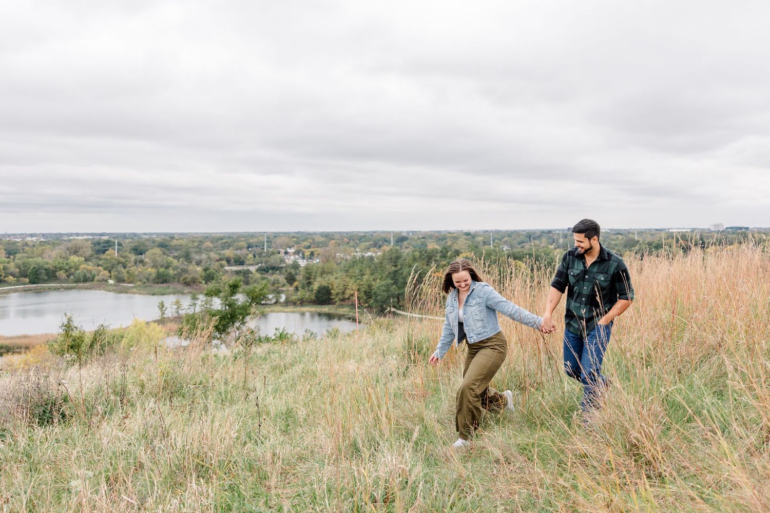 A smiling couple holds hands and walks up a grassy hillside overlooking a lake and trees under a cloudy sky.