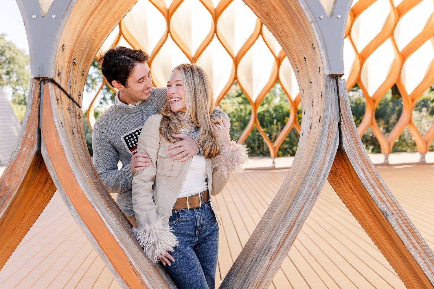 Happy couple embracing inside a decorative wooden lattice arch structure in a sunny park setting.
