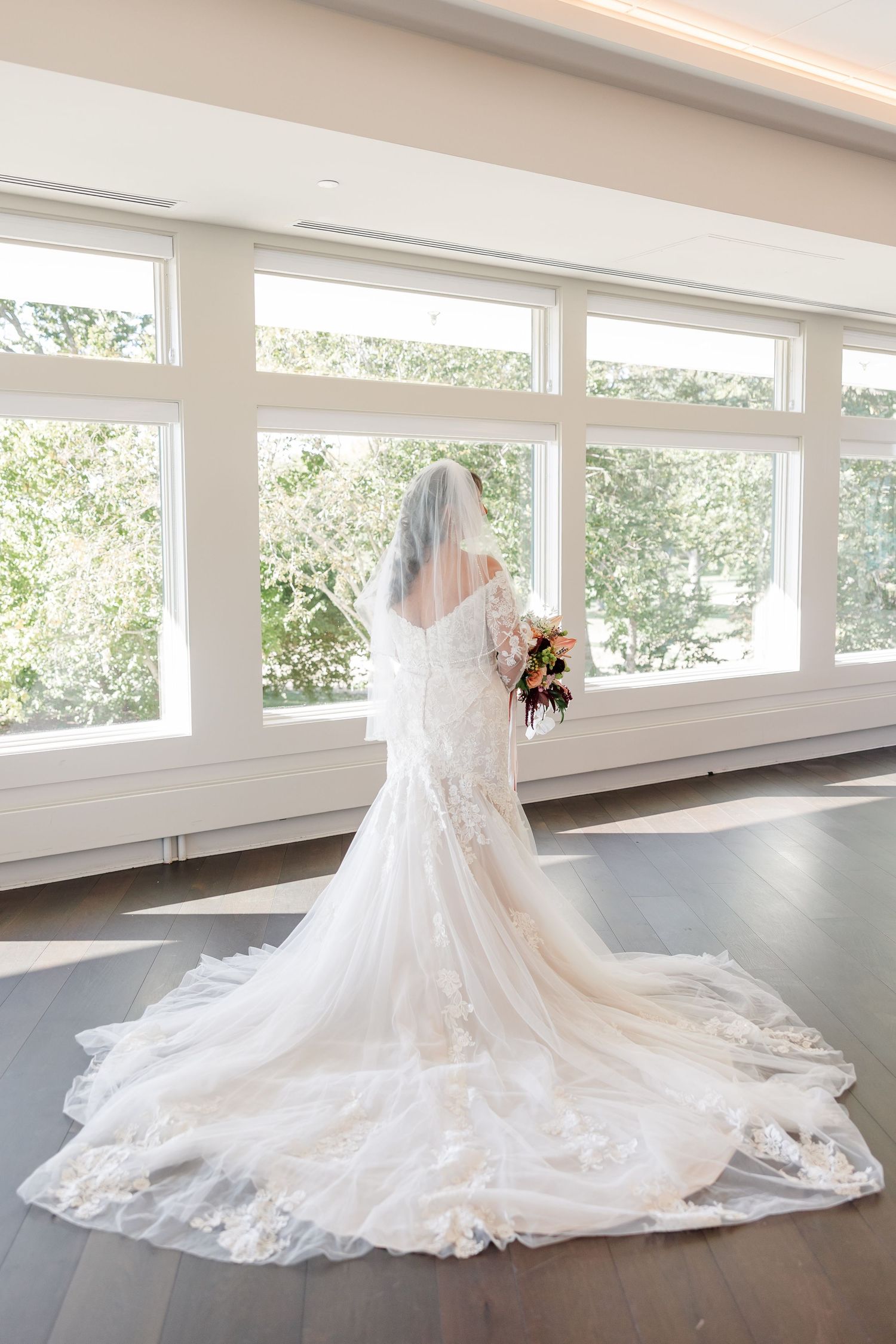 Bride in a lace wedding gown with a long train and veil holds a bouquet while facing large windows overlooking green trees.